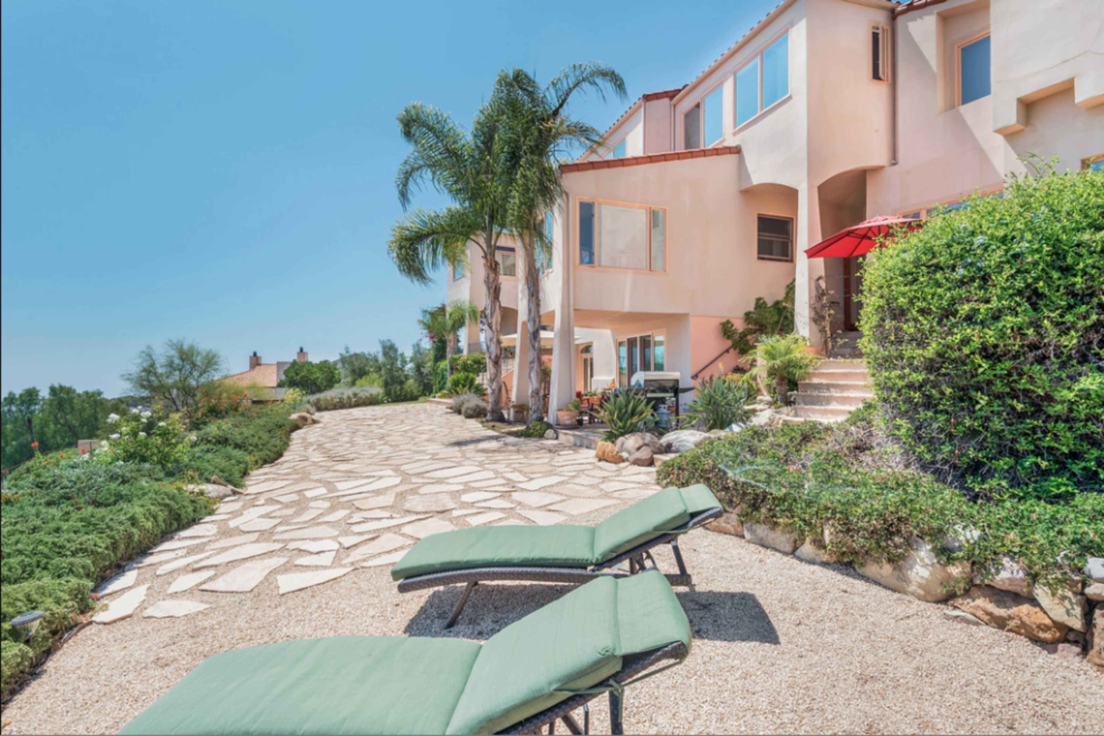 A pathway lined with palm trees and shrubs leads to a two-story stucco house with outdoor lounge chairs and an umbrella.