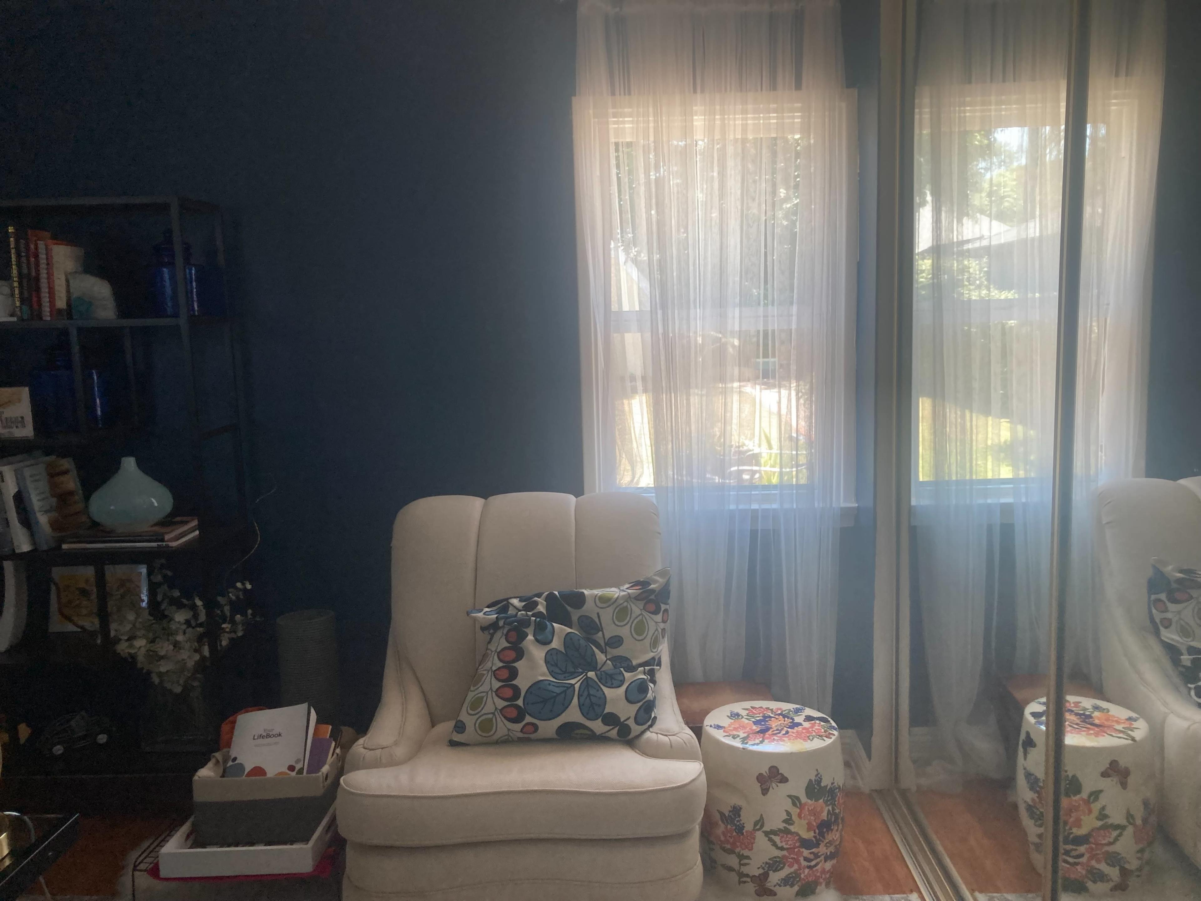 A cozy living room with two light-colored armchairs, a small decorative stool, and large windows dressed with sheer curtains allowing natural light to enter.