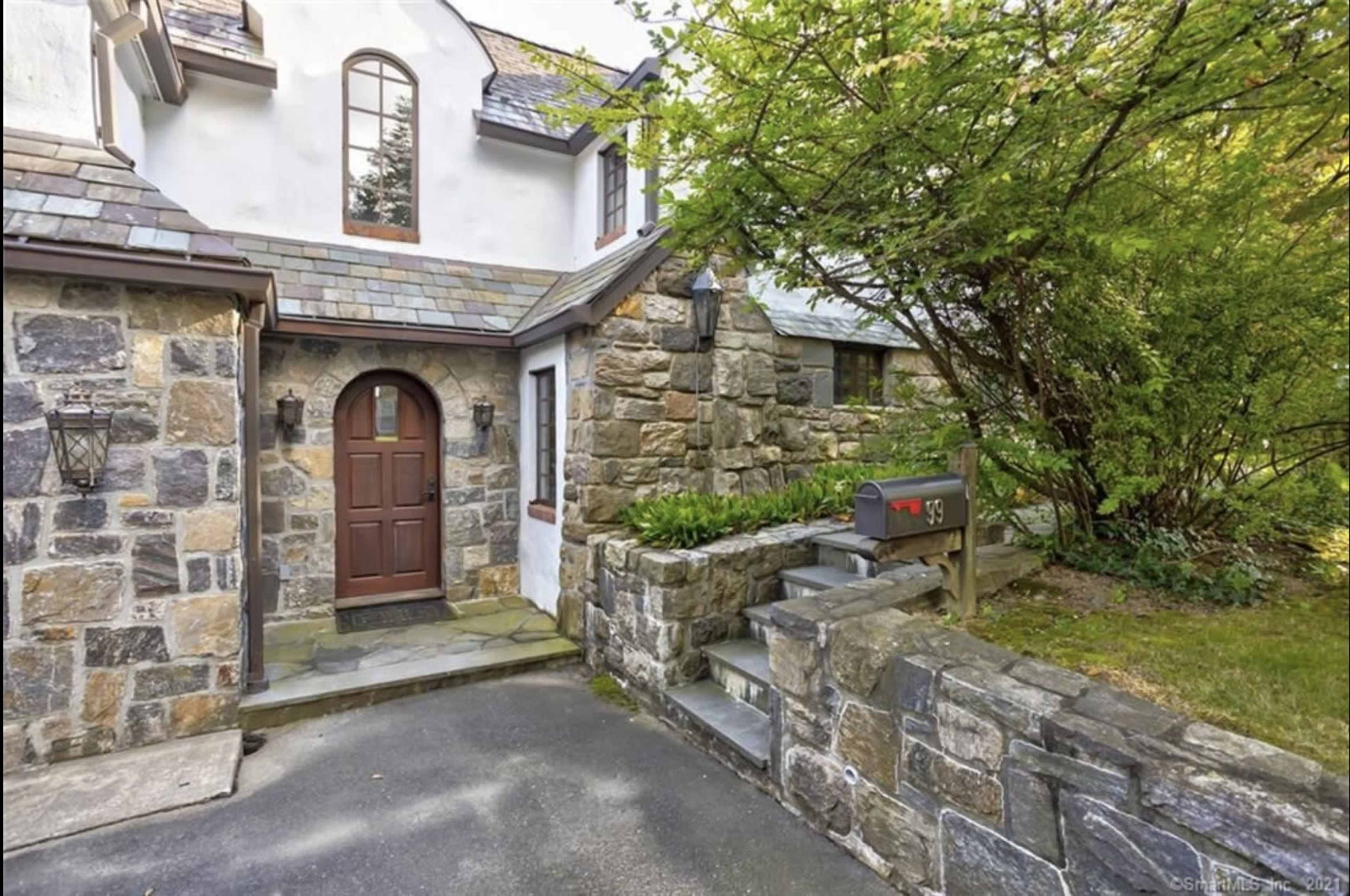 The entrance of a stone house with a wooden door, flanked by lanterns and surrounded by a landscaped garden.