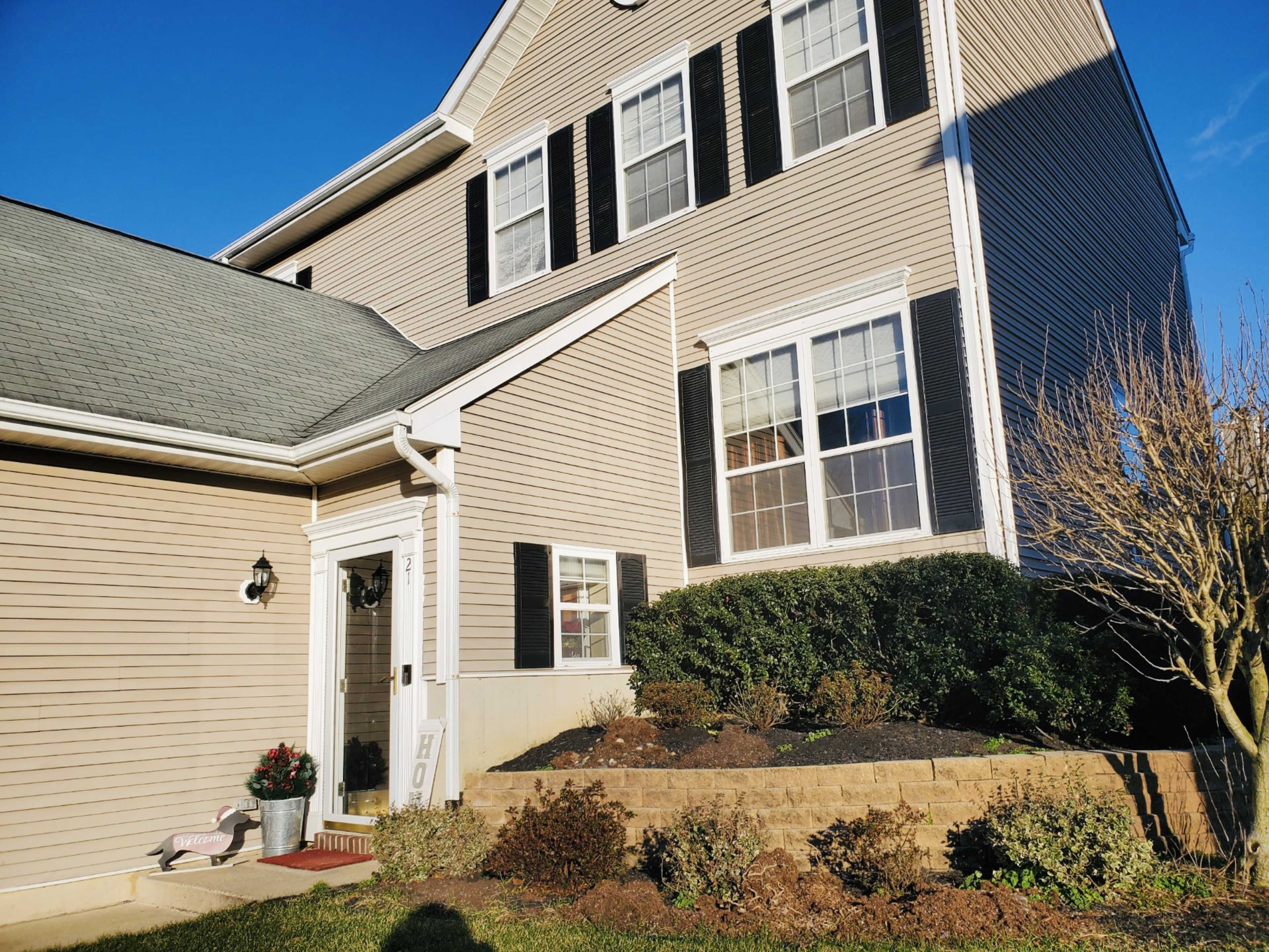 A two-story house with beige siding and black shutters features a front porch and landscaped garden.