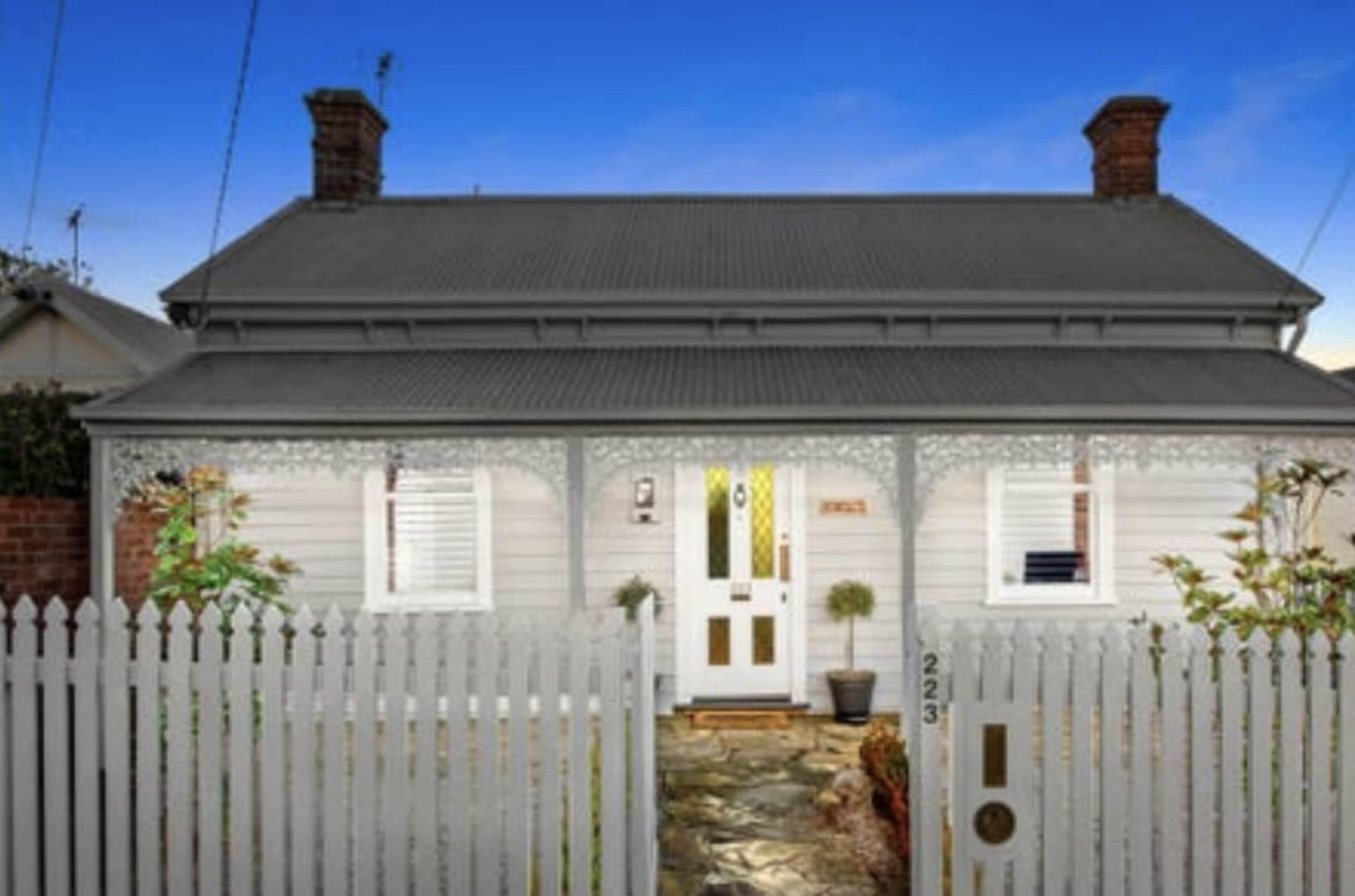 A single-story house with a gray roof, white wooden siding, and a picket fence, featuring a stone pathway leading to the front door.
