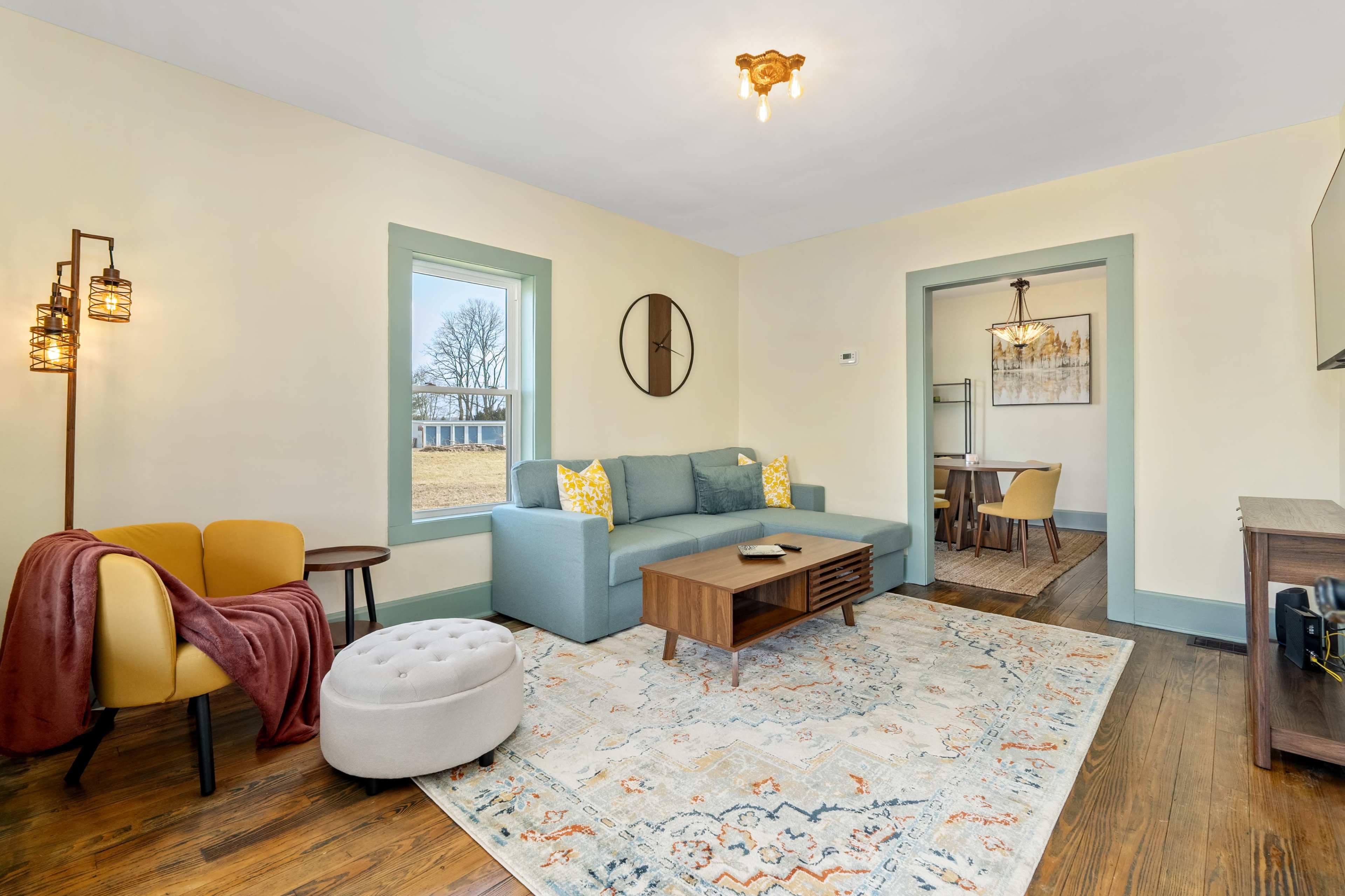 The image shows a cozy living room with a blue sofa, yellow accent pillows, a round ottoman, and a decorative rug.