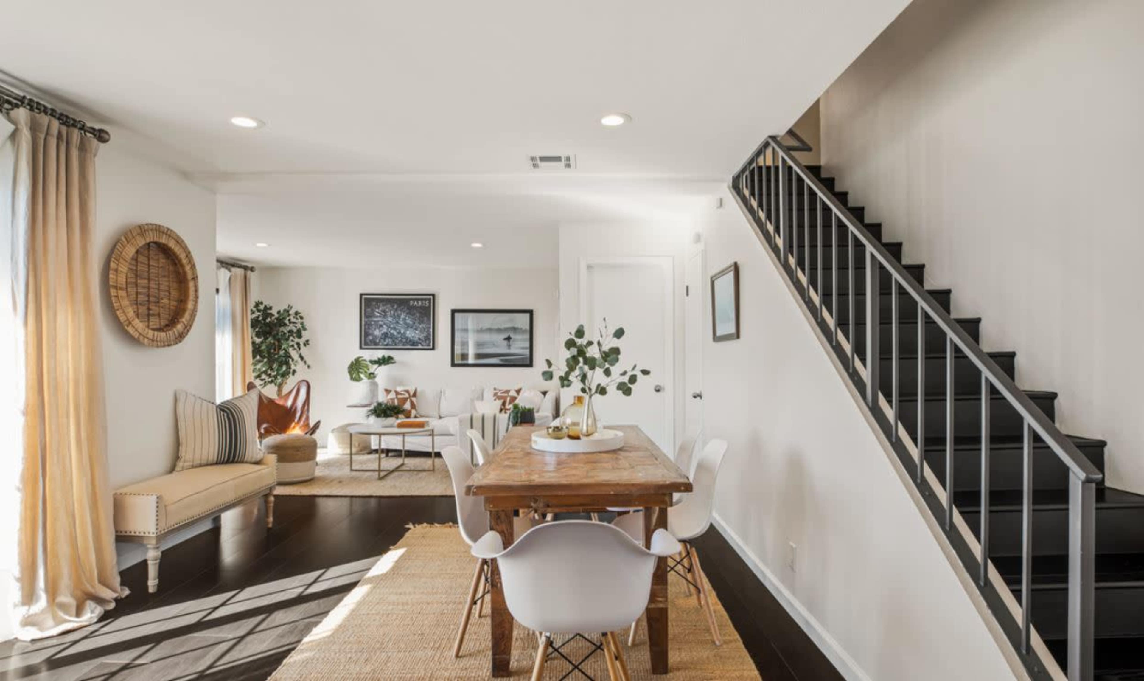 A bright dining area with a wooden table surrounded by white chairs, adjacent to a cozy living space featuring a sofa and a staircase to the right.