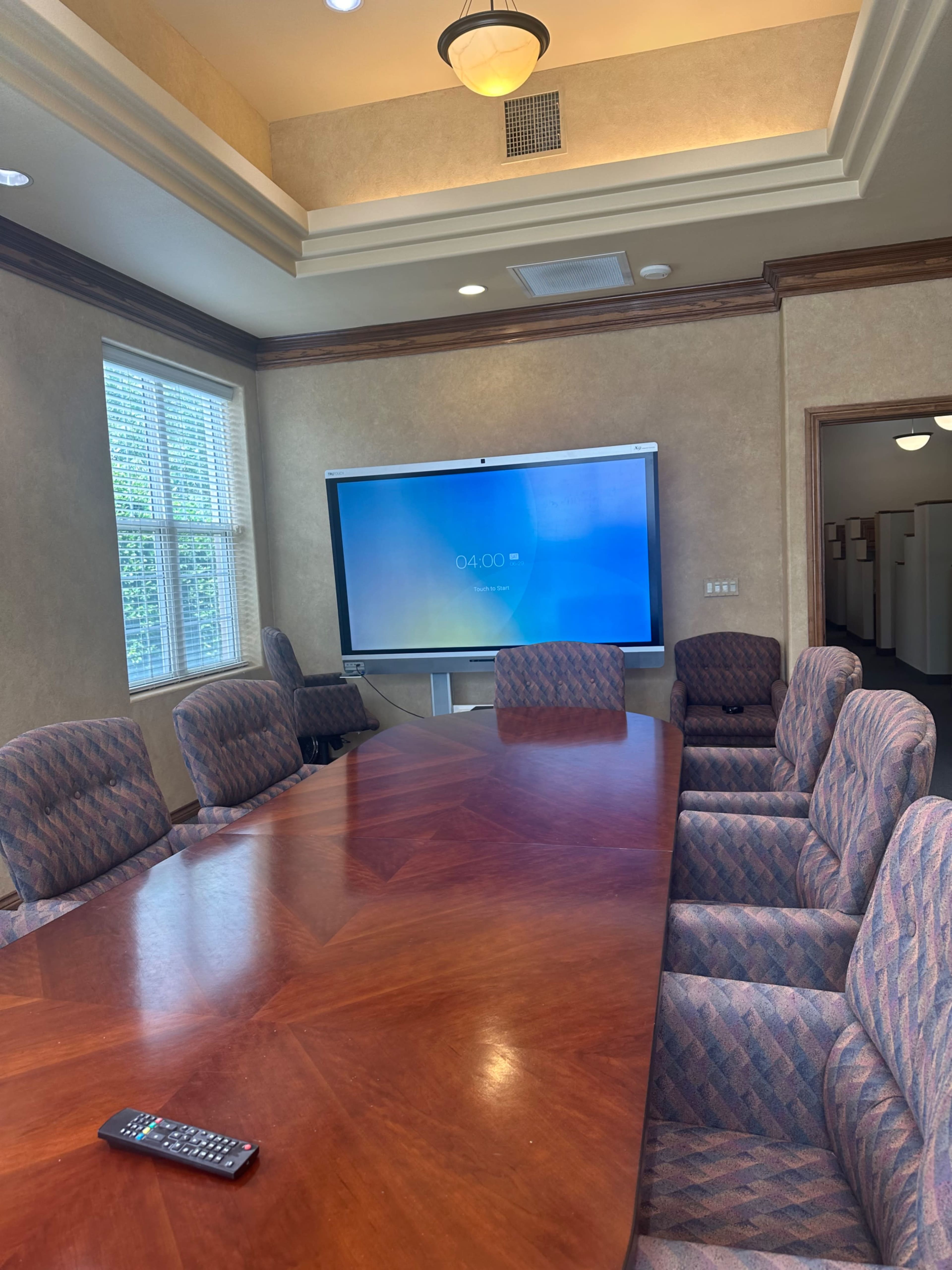 A conference room features a large wooden table surrounded by upholstered chairs and a wall-mounted display screen.