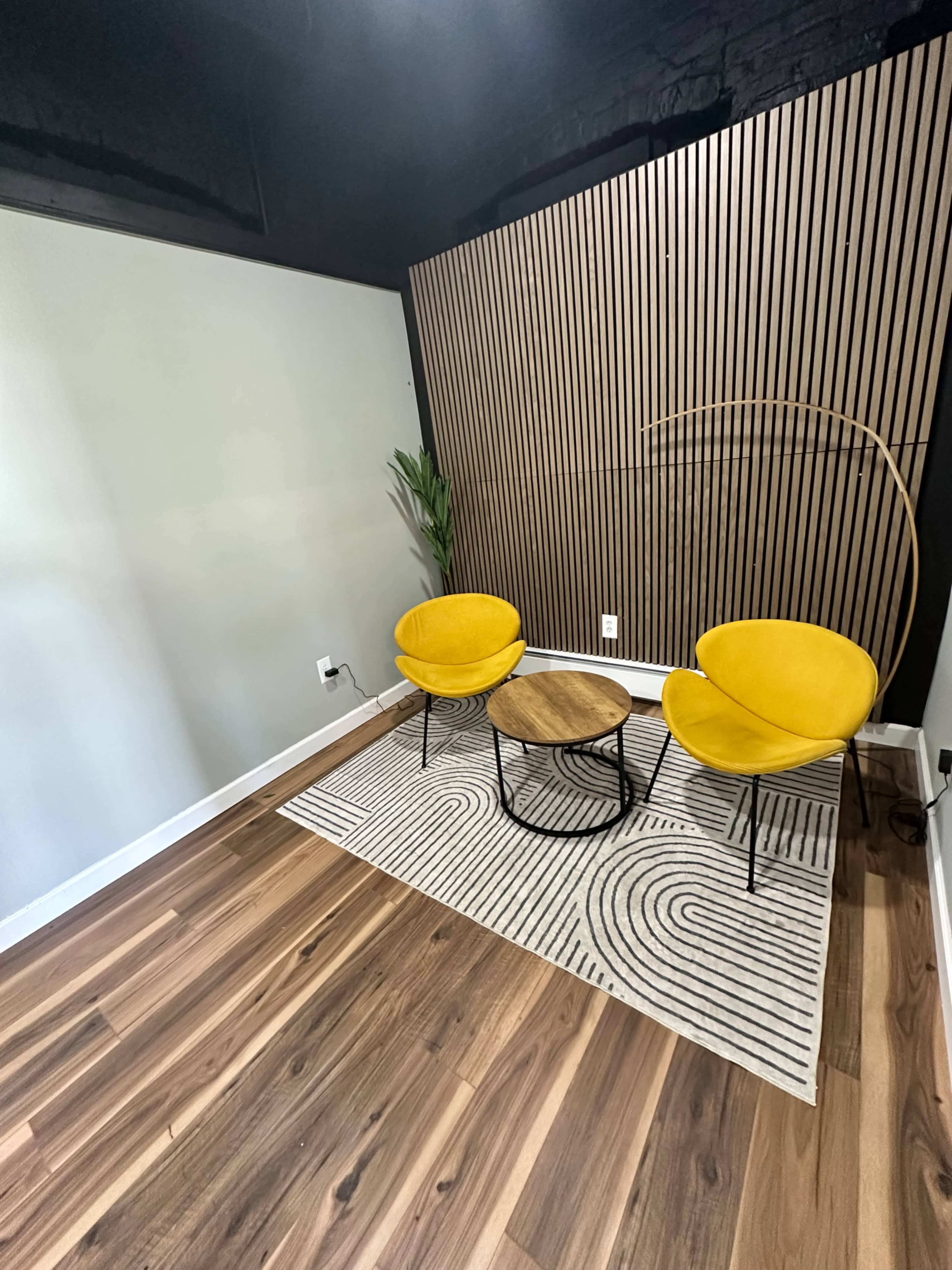 The image shows a minimalist interior with two yellow chairs, a small round wooden table on a patterned rug, and a wall featuring vertical wooden slats.