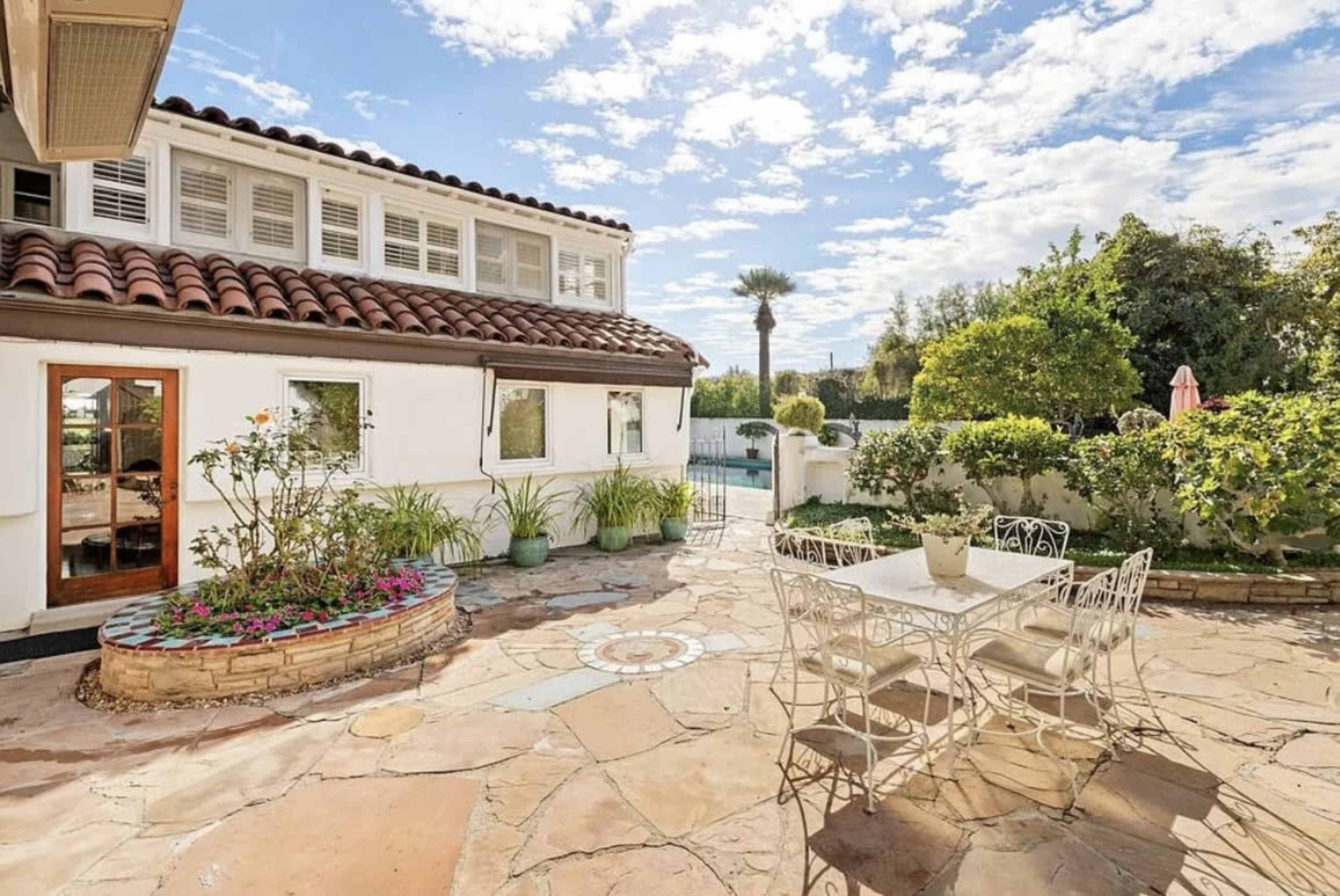 A stone patio features a dining table surrounded by chairs, with potted plants and a pool visible in the background.