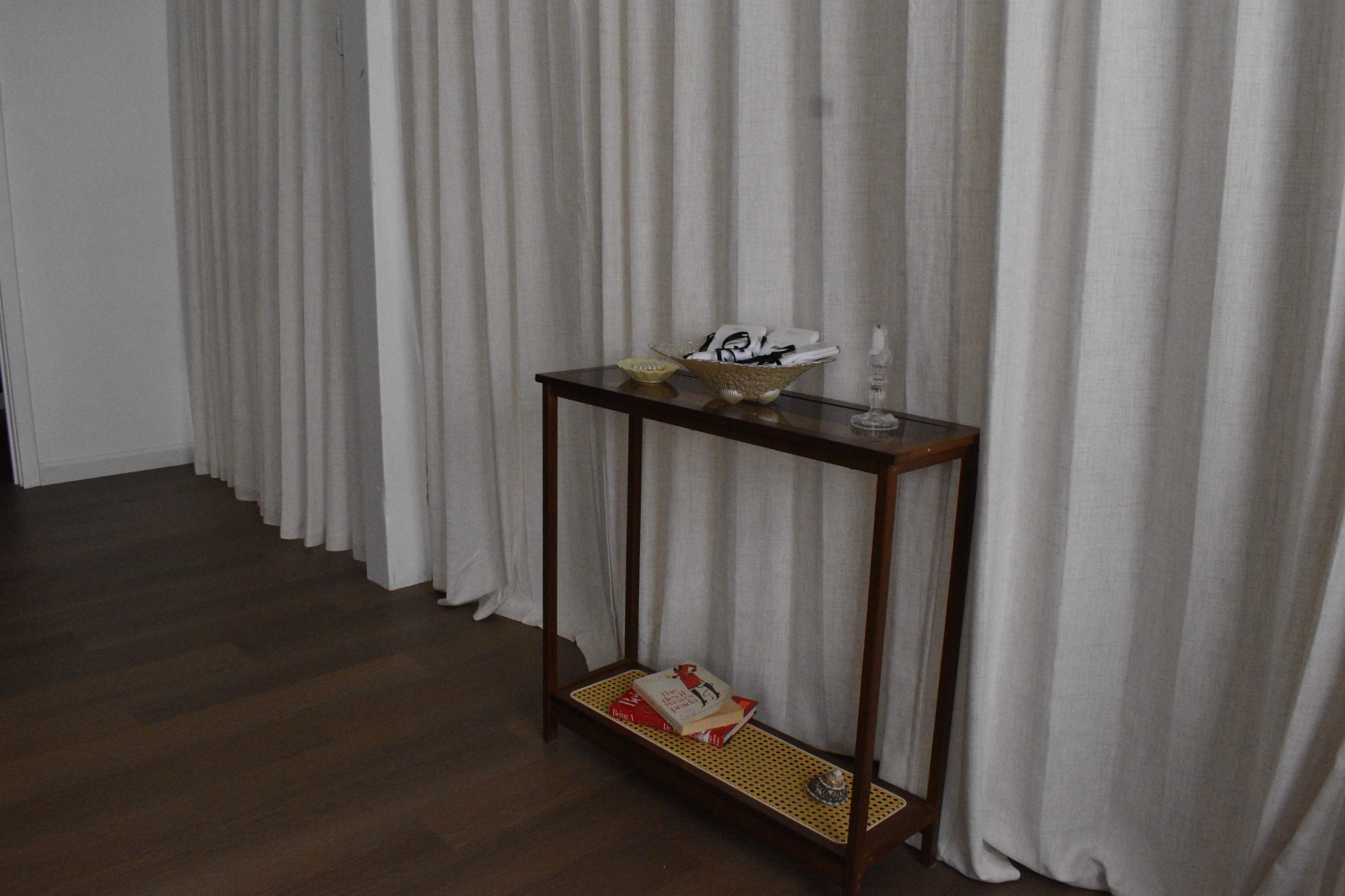 A wooden table with a glass top and several decorative items sits against a wall, next to long, light-colored curtains.