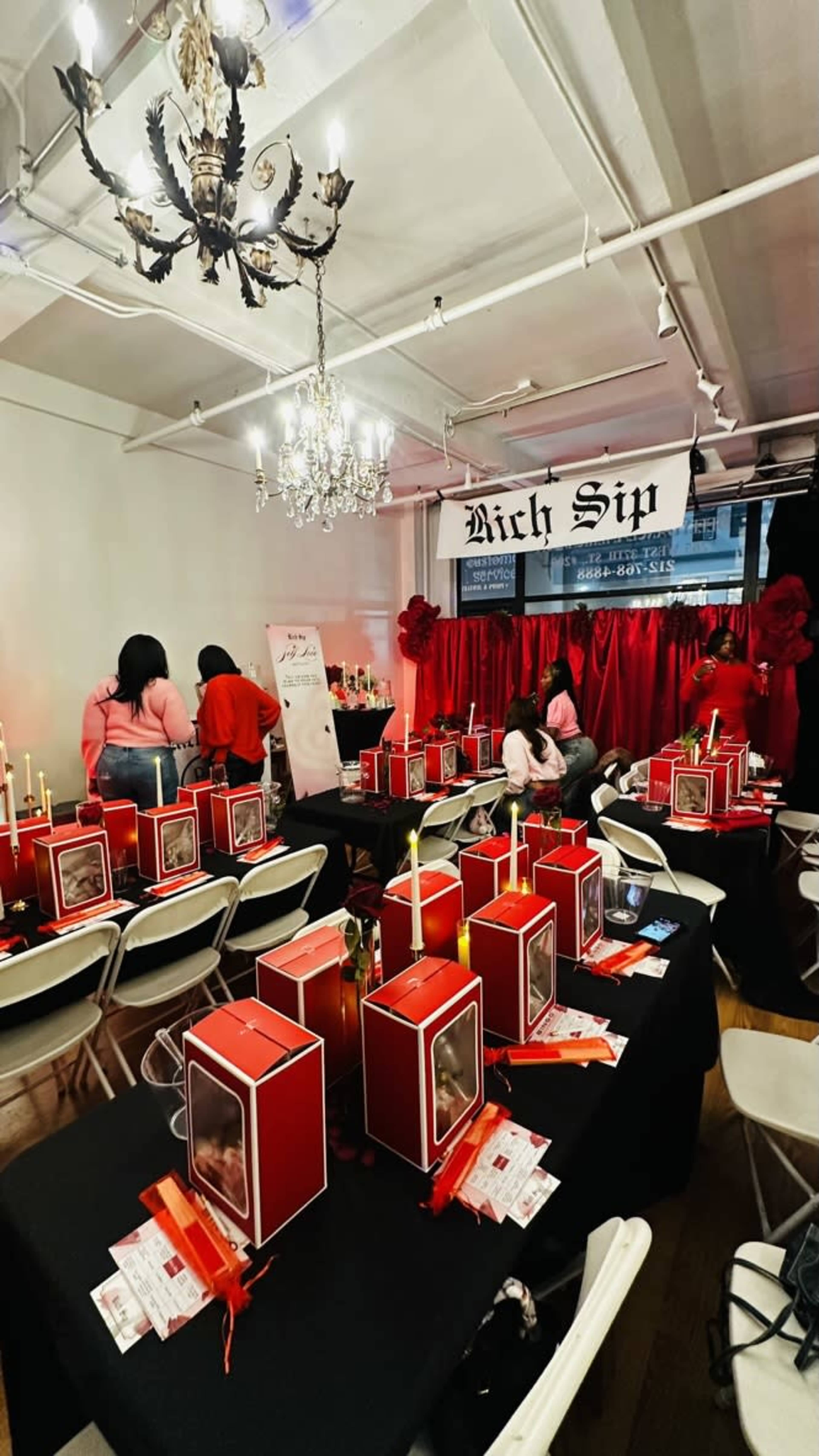 The scene shows a decorated indoor space with several tables set up, each adorned with red boxes and candles, while people are interacting in the background.