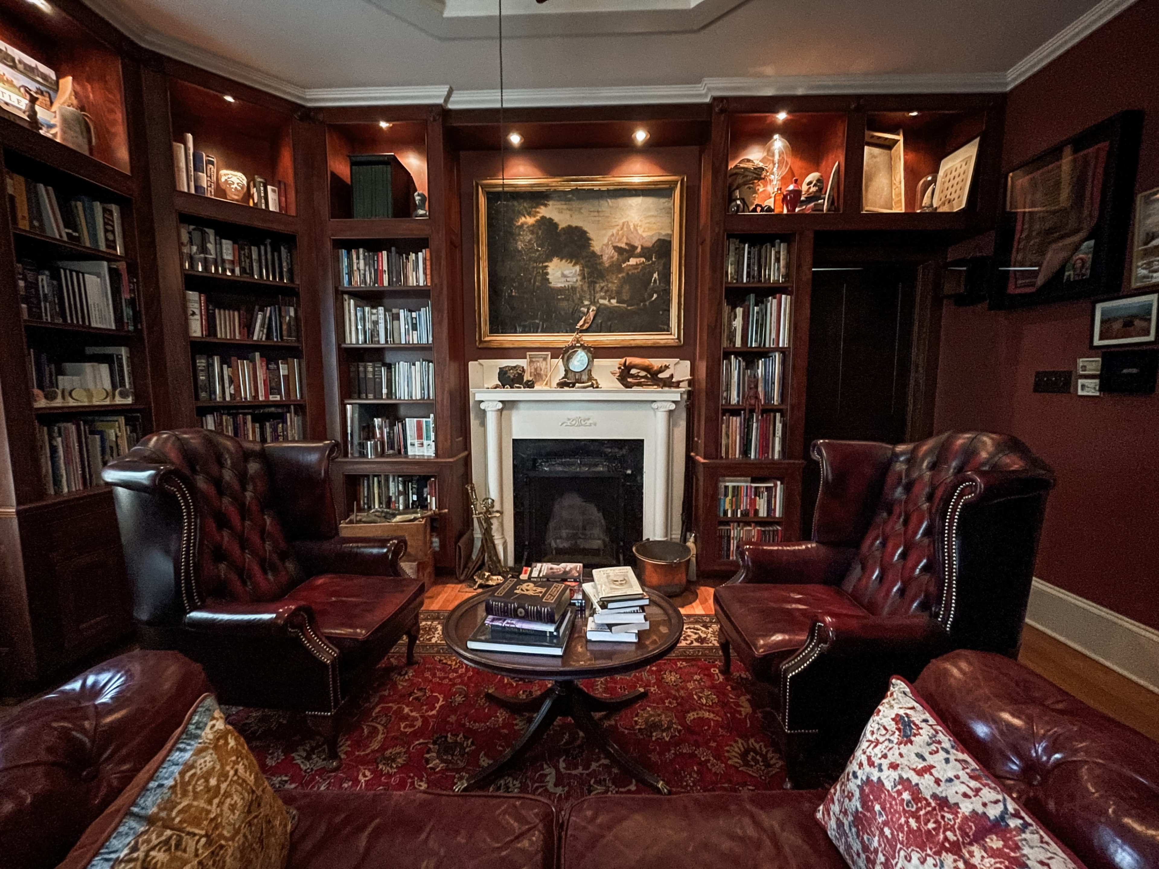 A cozy library featuring two leather armchairs facing a small circular table, surrounded by bookshelves and a fireplace with a large painting above it.