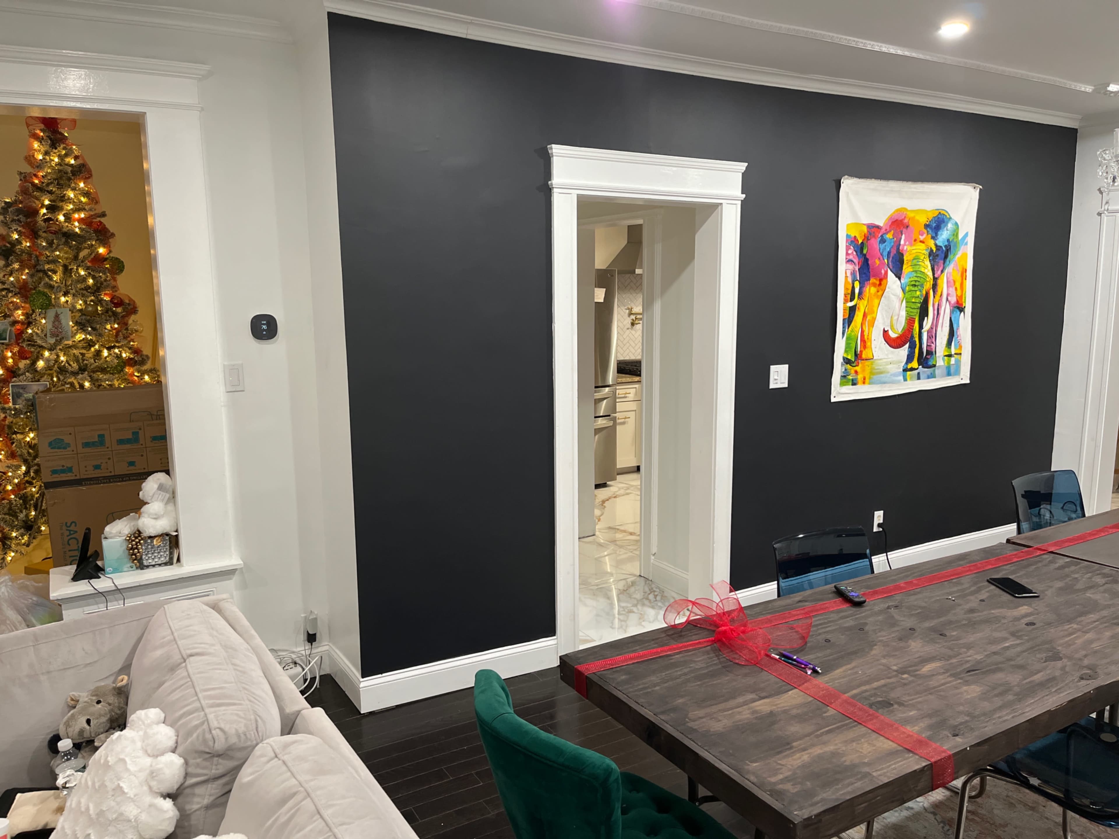 The image shows a dining area with a dark wall featuring an elephant painting, a dining table in the center, and a Christmas tree visible in the background.