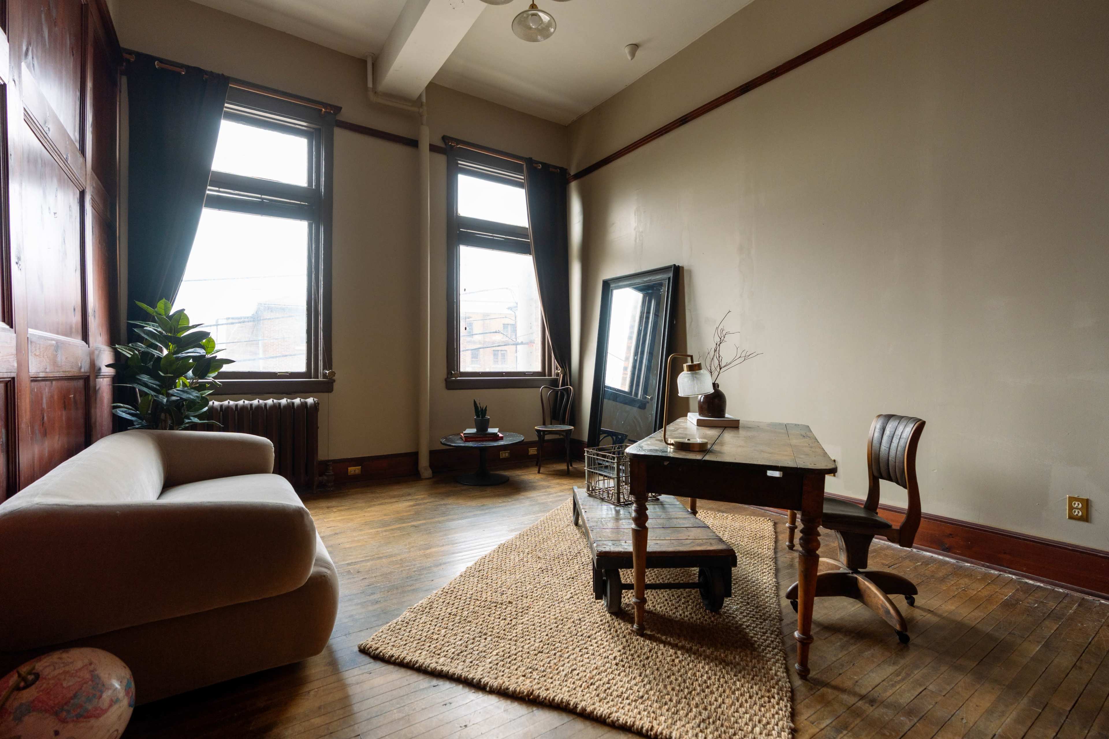 The image shows a spacious room with hardwood floors, featuring a sofa, a wooden table and chair, and large windows allowing natural light to enter.