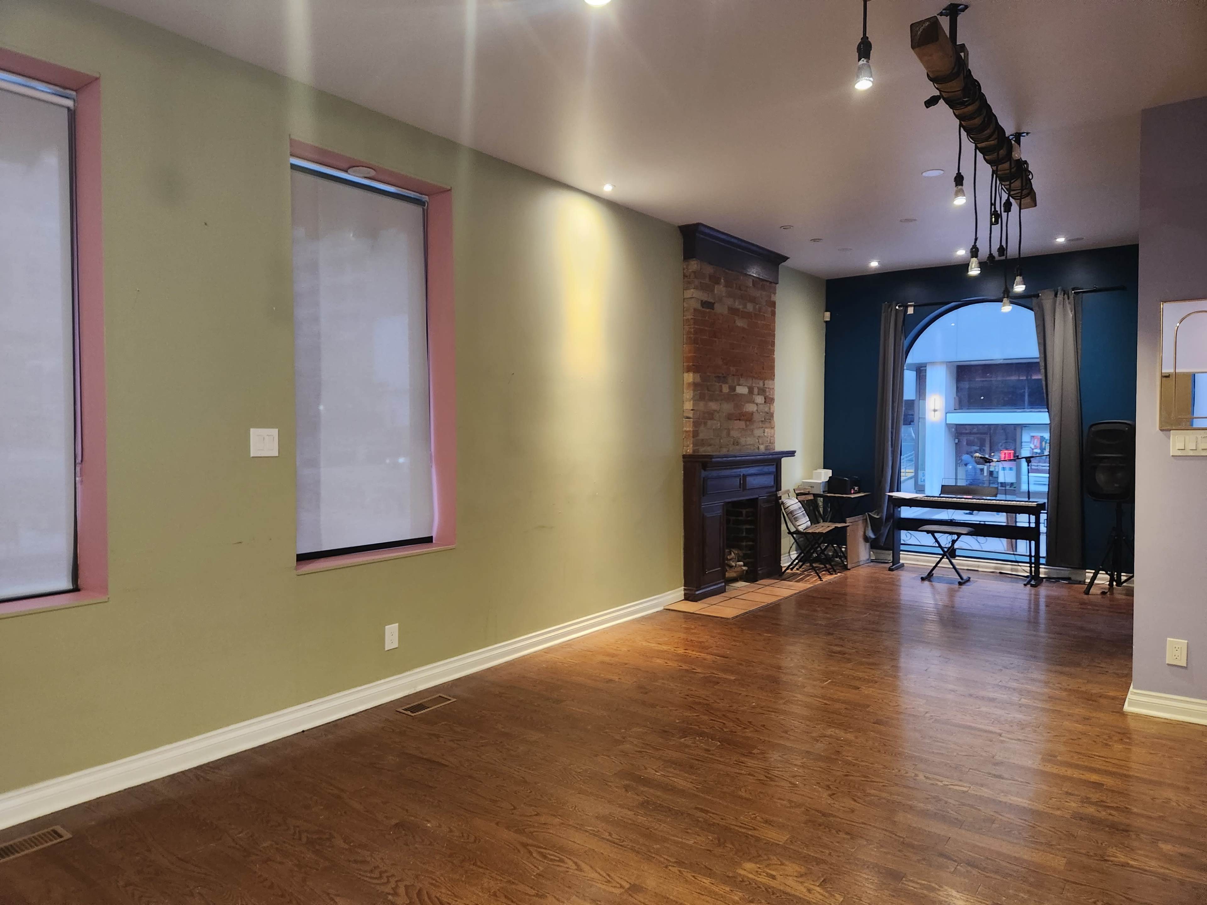 The room features hardwood flooring, two windows with shades, a brick fireplace, and a small table and chair set in an adjacent area.