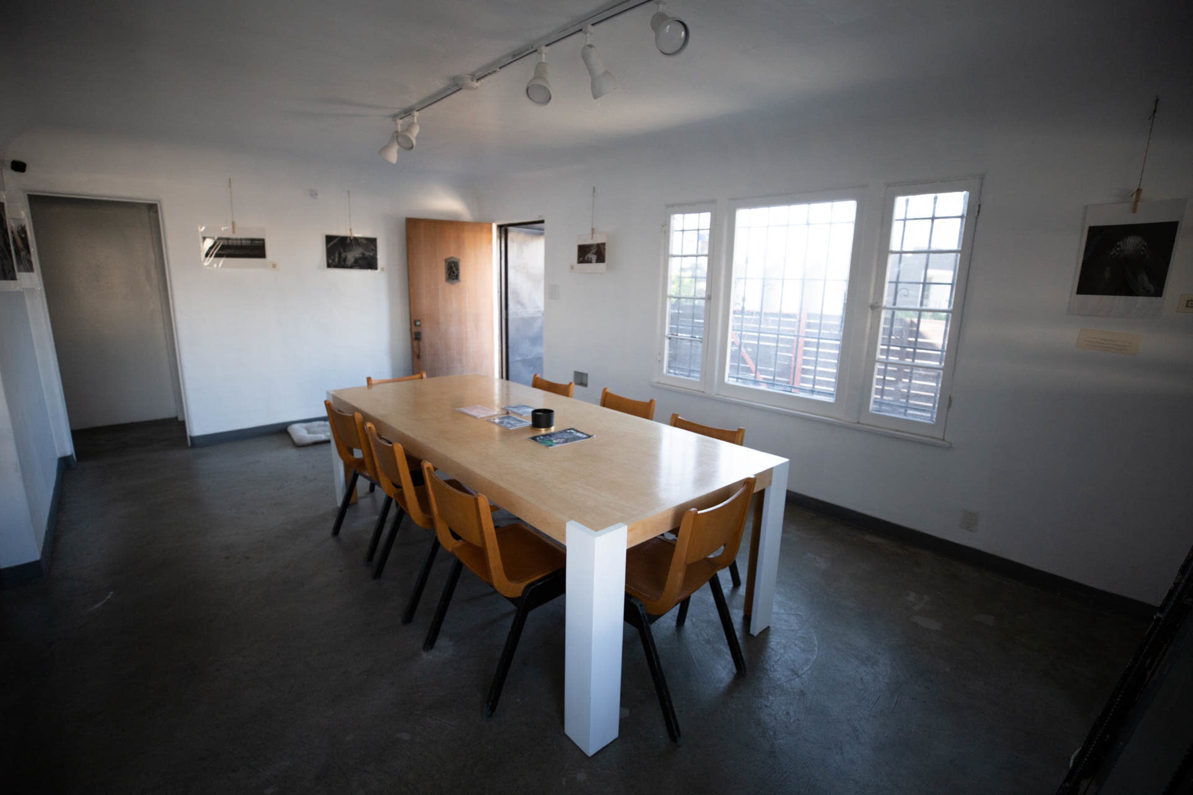 A long wooden table with eight chairs is positioned in a well-lit room featuring large windows and minimal decorations.
