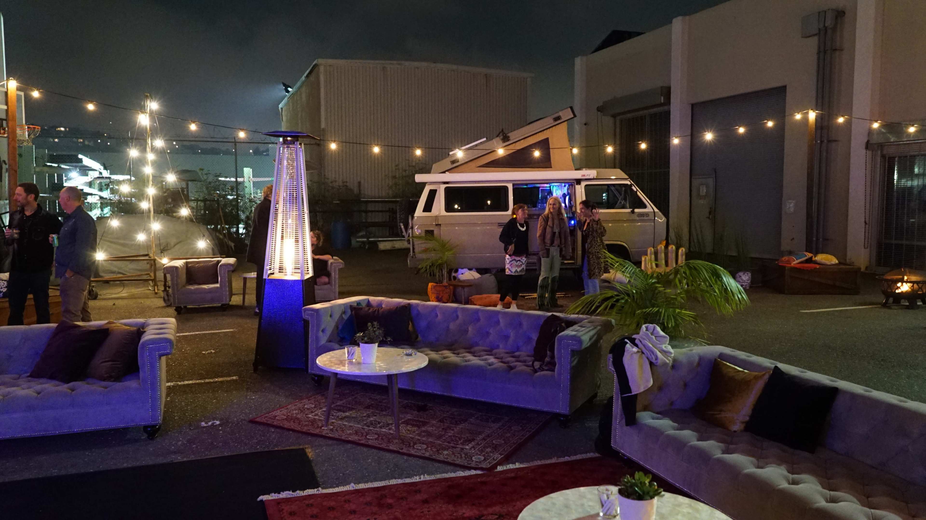 A cozy outdoor event space features chic sofas, string lights, a vintage camper van, and guests socializing amidst decorative elements like plants and a fire pit.