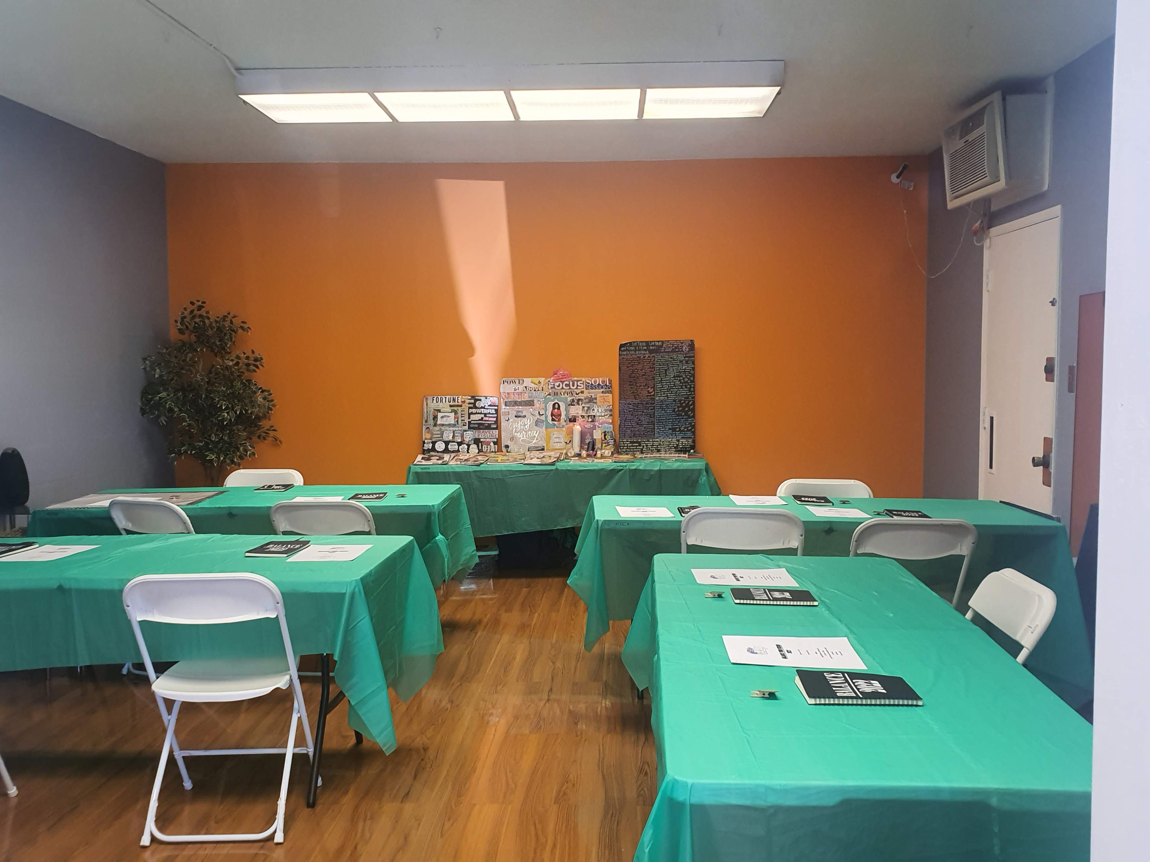 The image shows a room set up with several green tables covered with white chairs, facing a display table against an orange wall.