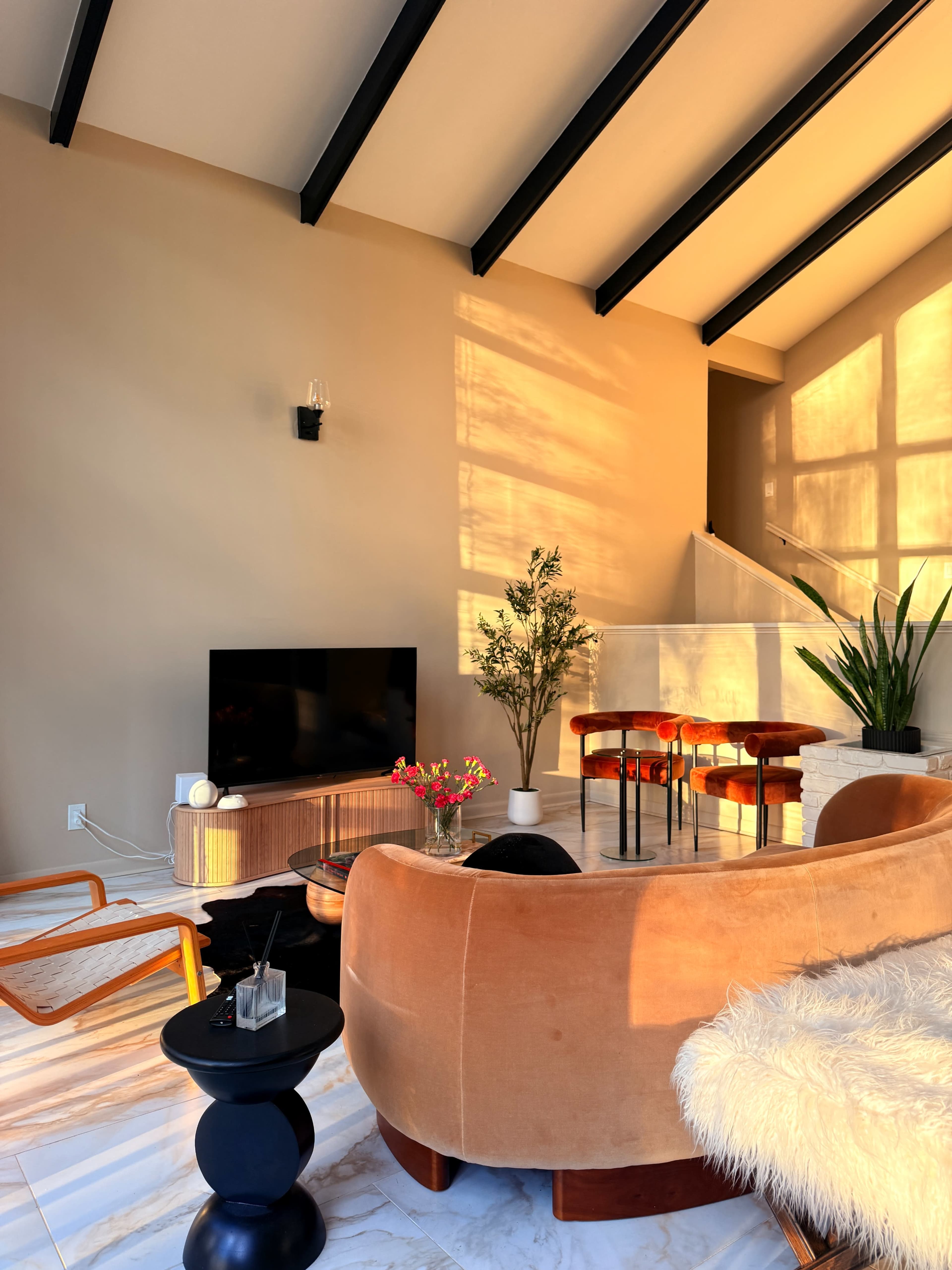 A modern living room features a curved orange sofa, a black television on a wooden stand, a small table, and a dining setup with four stools, illuminated by warm sunlight through large windows.