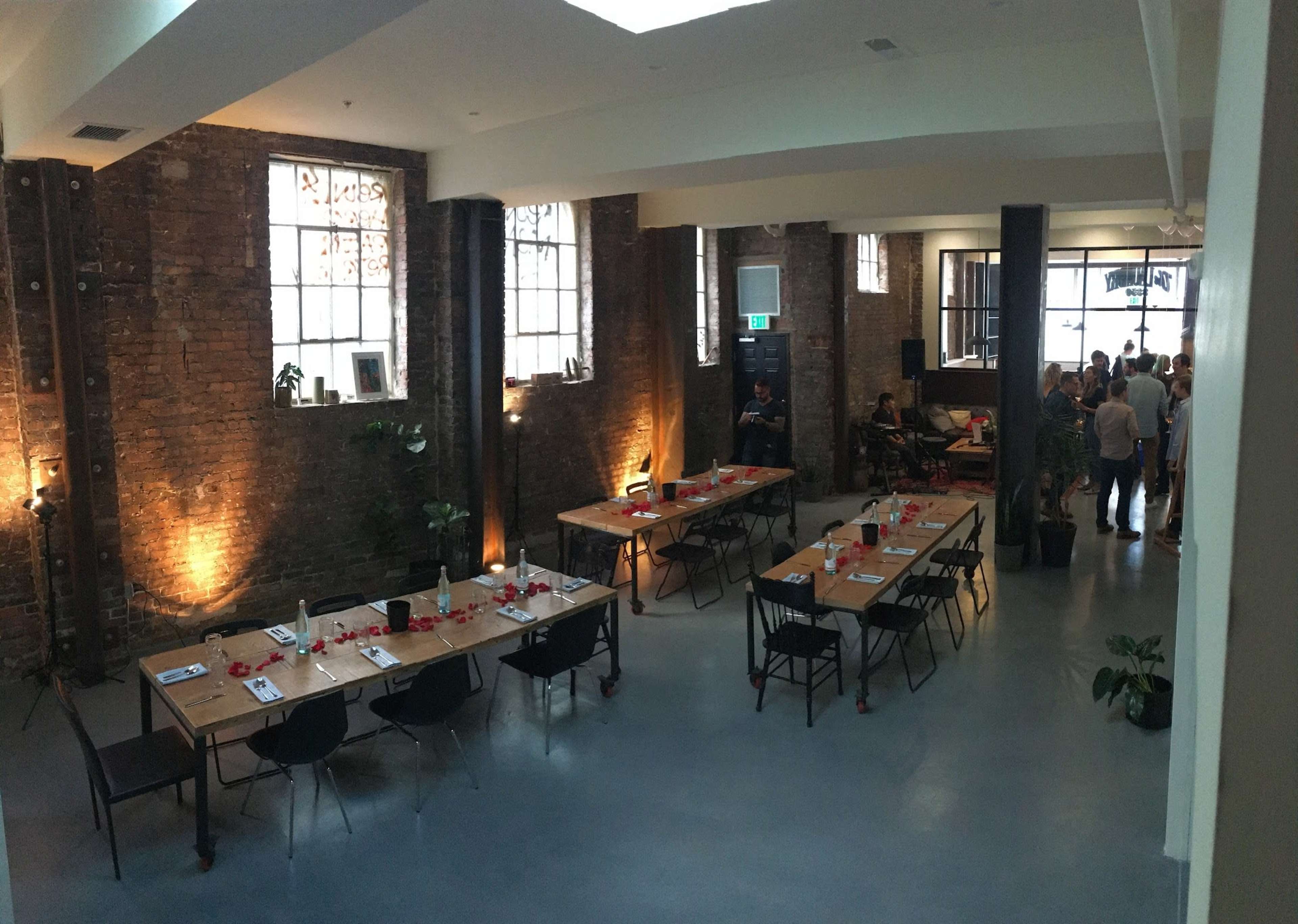 The image shows a spacious, industrial-style room with large windows, featuring multiple long tables arranged for a gathering and people socializing in the background.