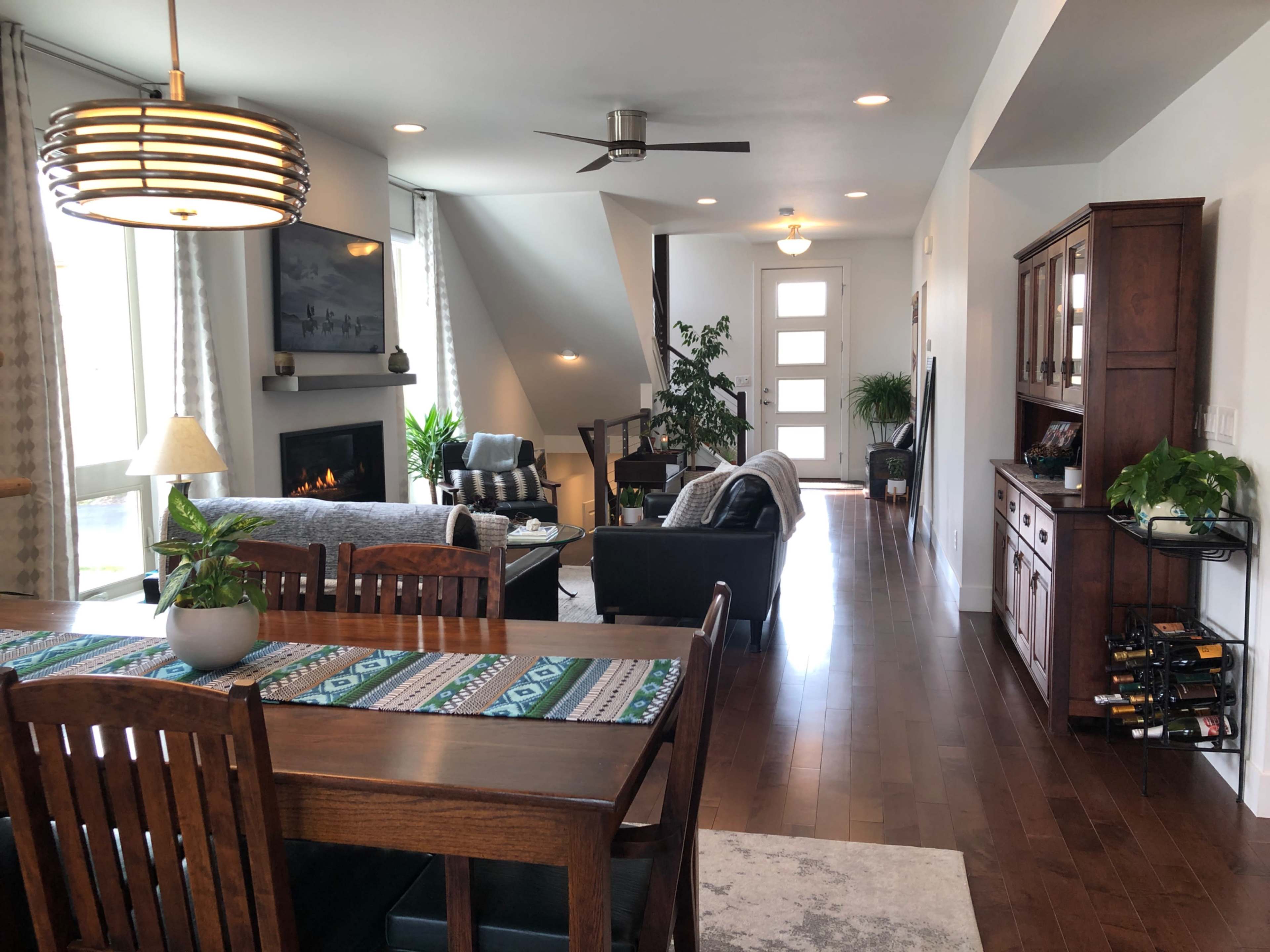 The image shows a modern living space featuring a dining area with a wooden table, chairs, and a decorative table runner, leading into a lounge area styled with a sofa, armchairs, and a fireplace, with natural light streaming through large windows.