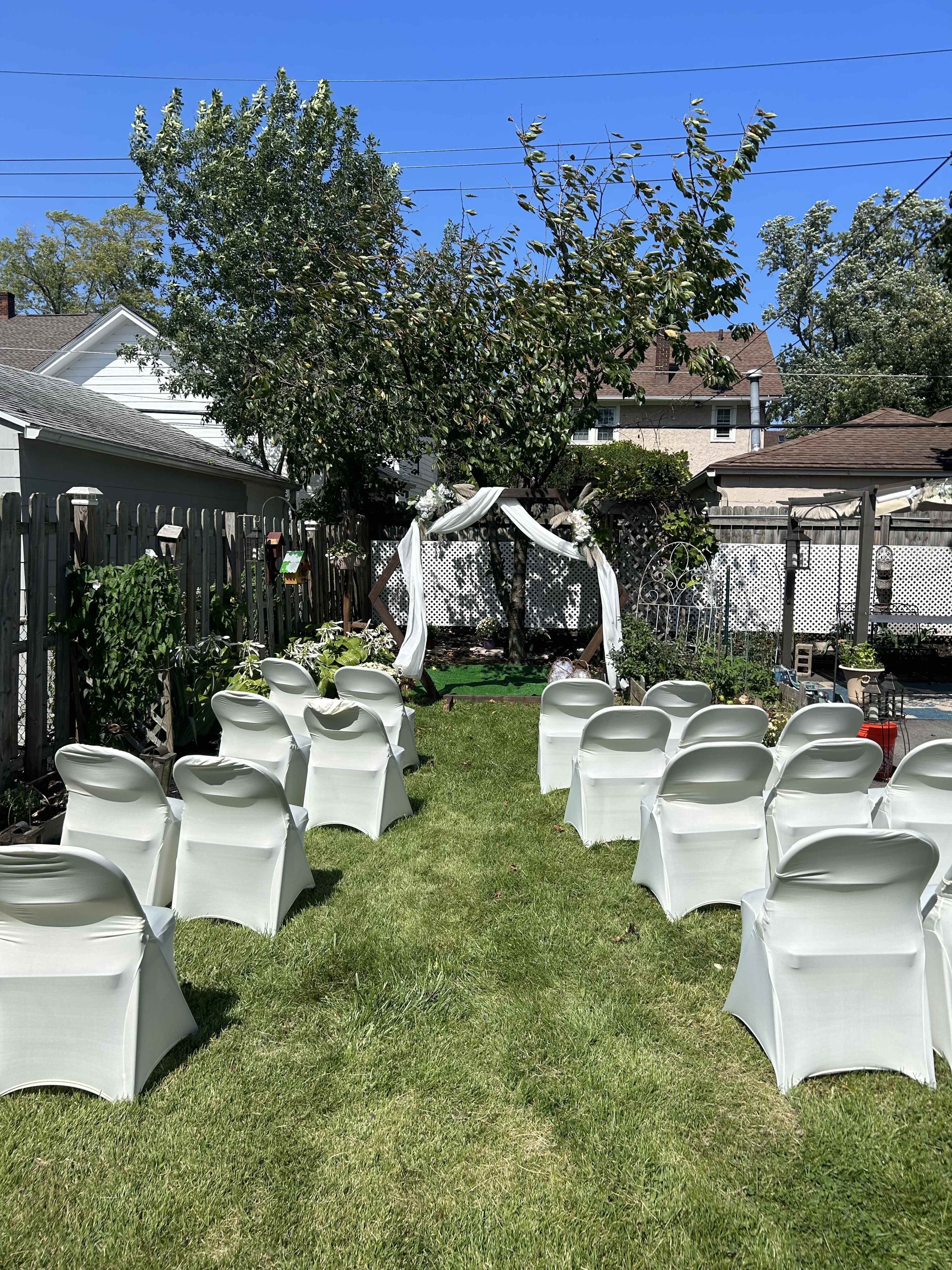 A backyard is set up with white chairs facing a decorative arch under a tree for an outdoor event.
