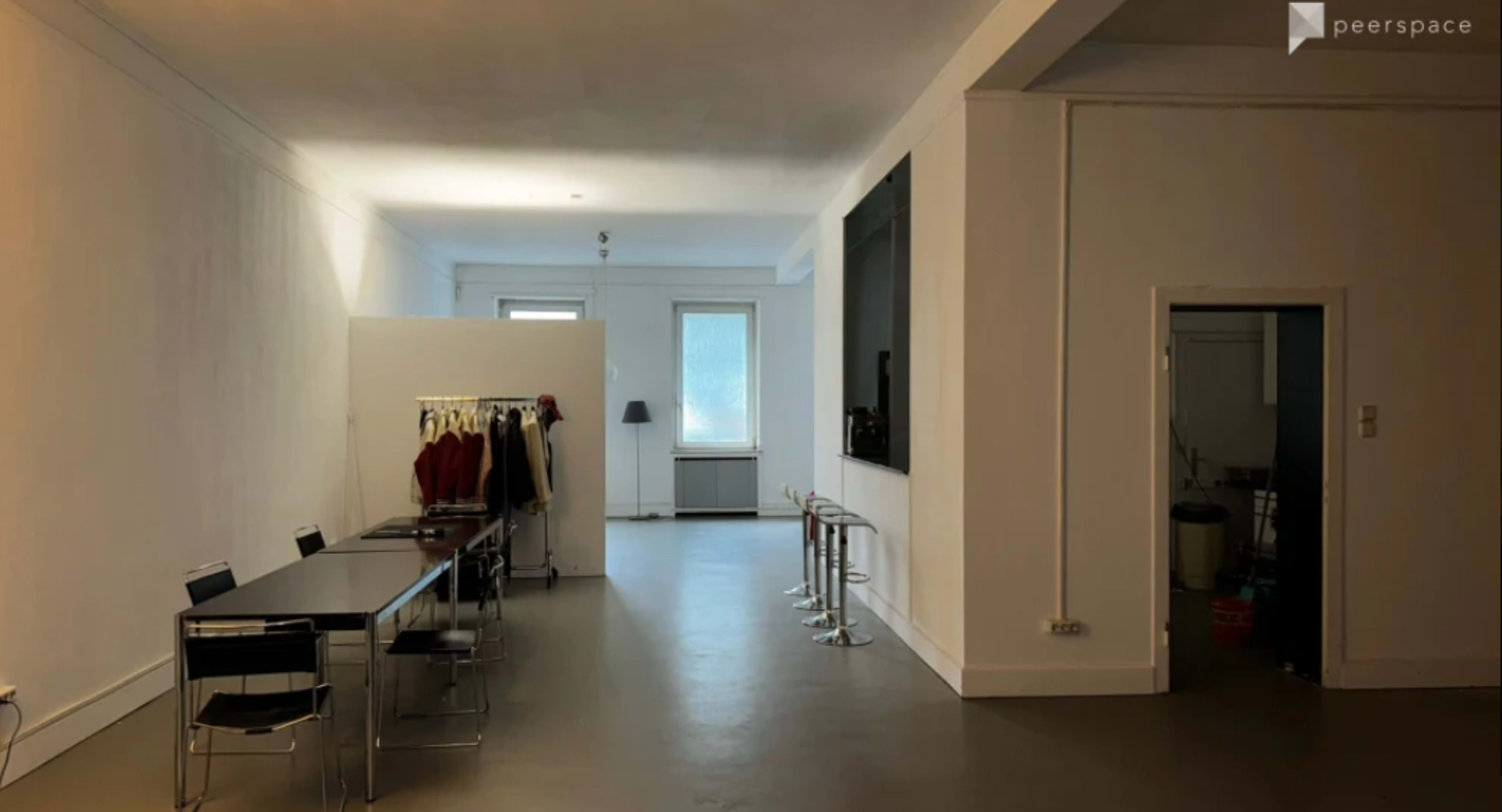The image shows a spacious, minimalist room featuring a long dining table with chairs, a clothing rack against one wall, and a set of sleek bar stools near a small kitchenette area.