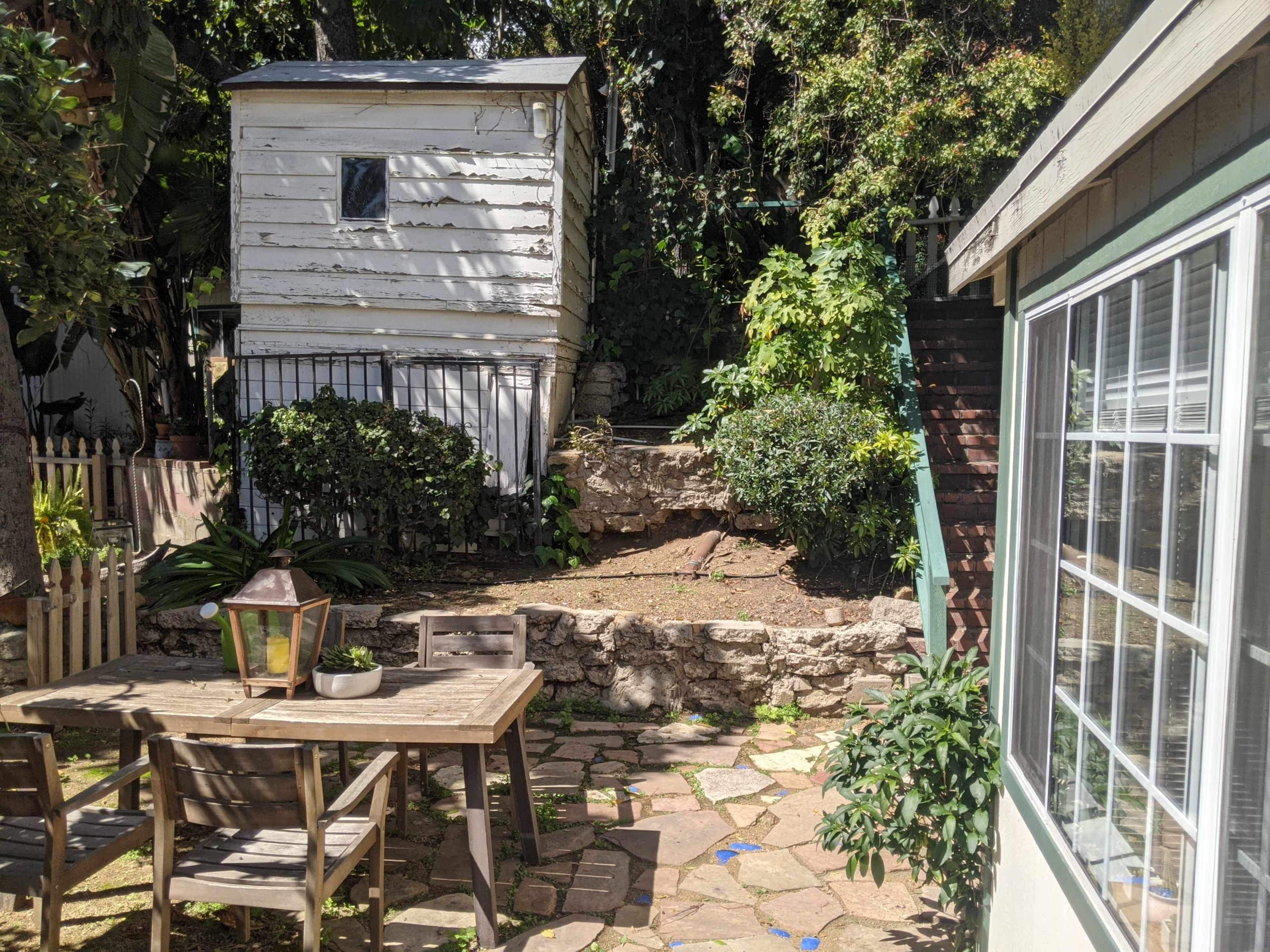 The image shows a small outdoor area with a wooden table and chairs, a weathered shed, a stone wall, and stairs leading up to a higher level.