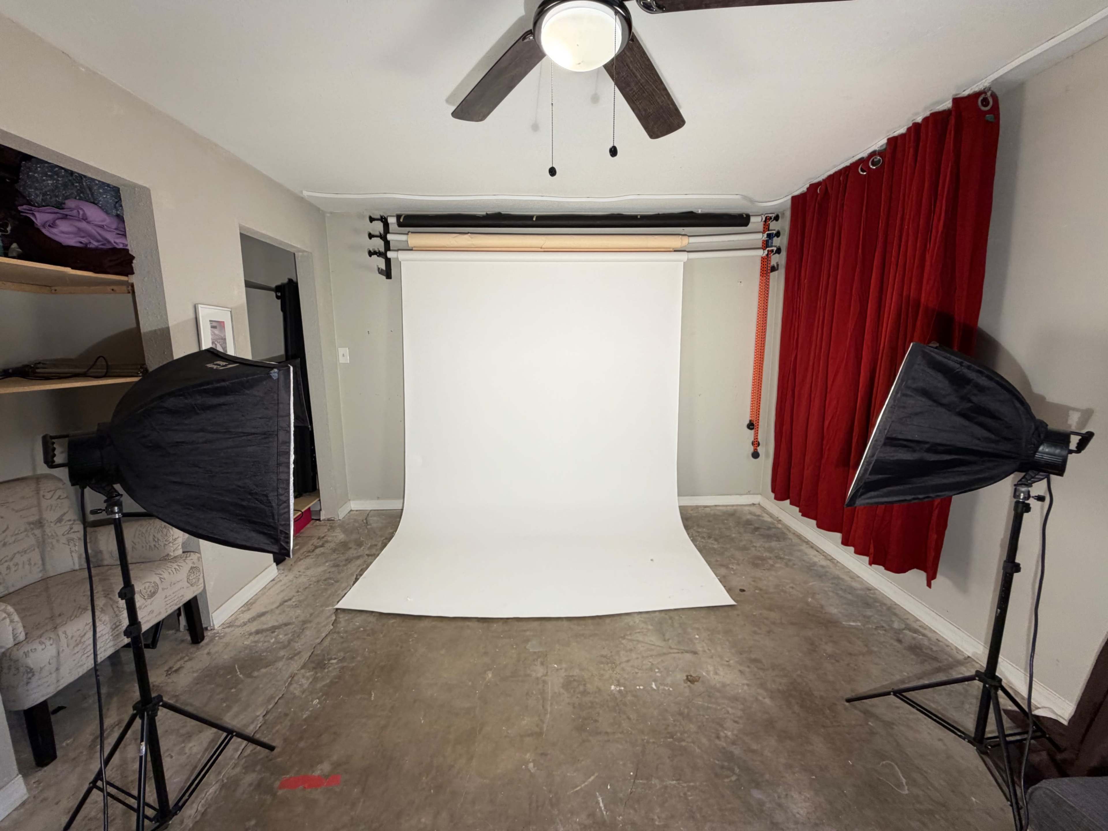 The image shows a photography studio featuring a plain white backdrop, two softbox lights on tripods, and red curtains on one side.