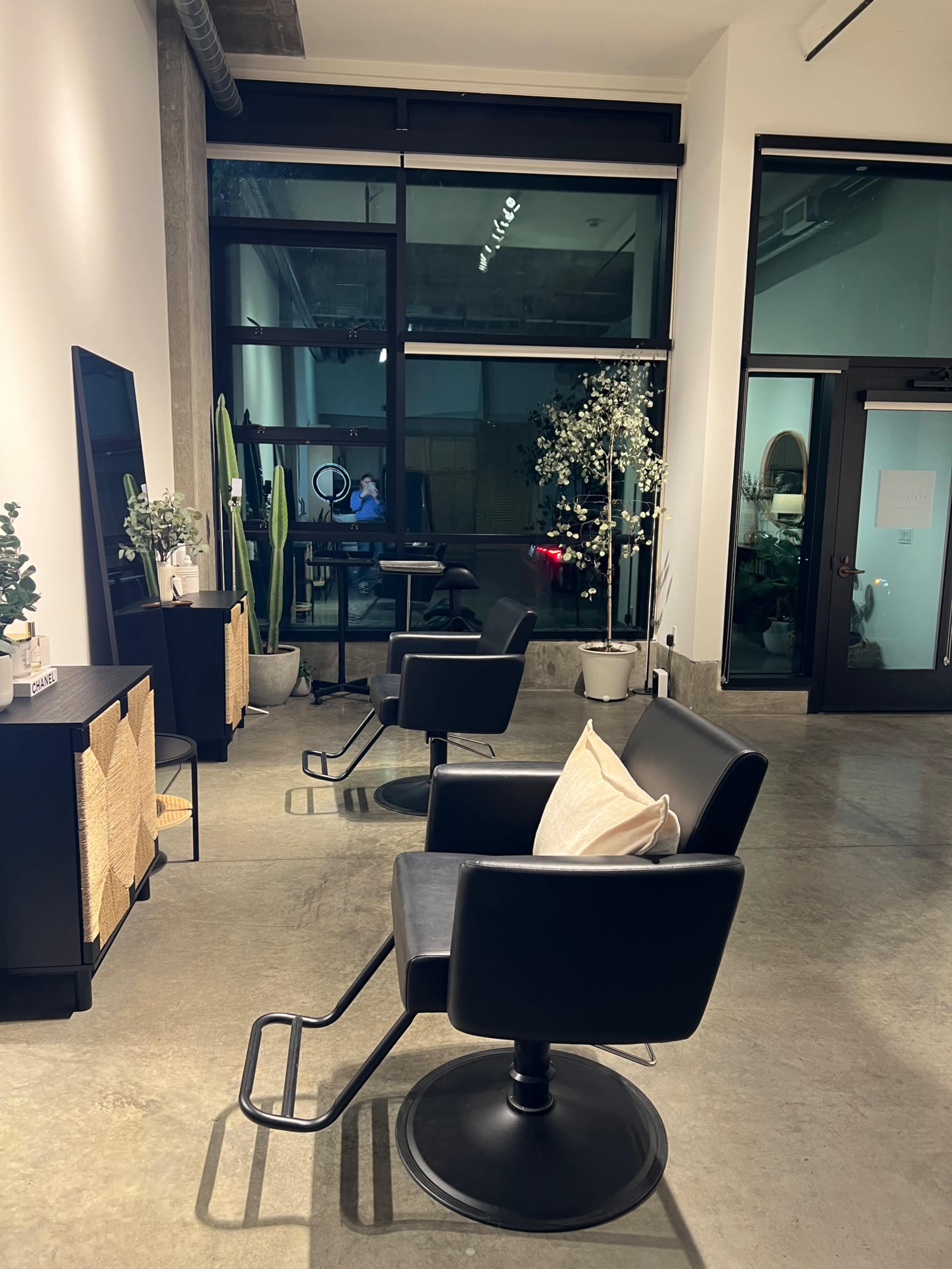 The image shows a modern salon interior with two black styling chairs and plants, illuminated by soft lighting.