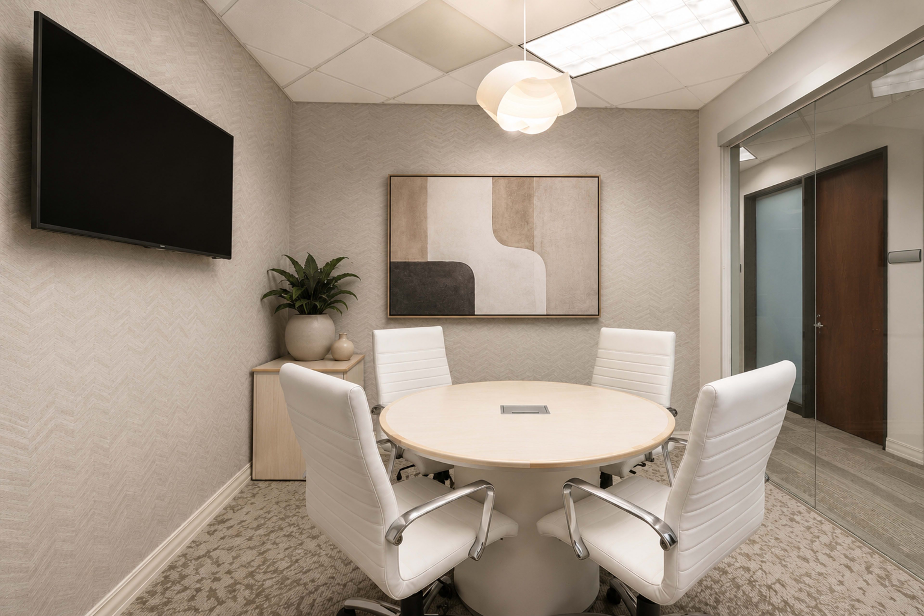 A modern conference room features a round table surrounded by four white chairs, a large wall-mounted TV, and a piece of abstract art on the wall.