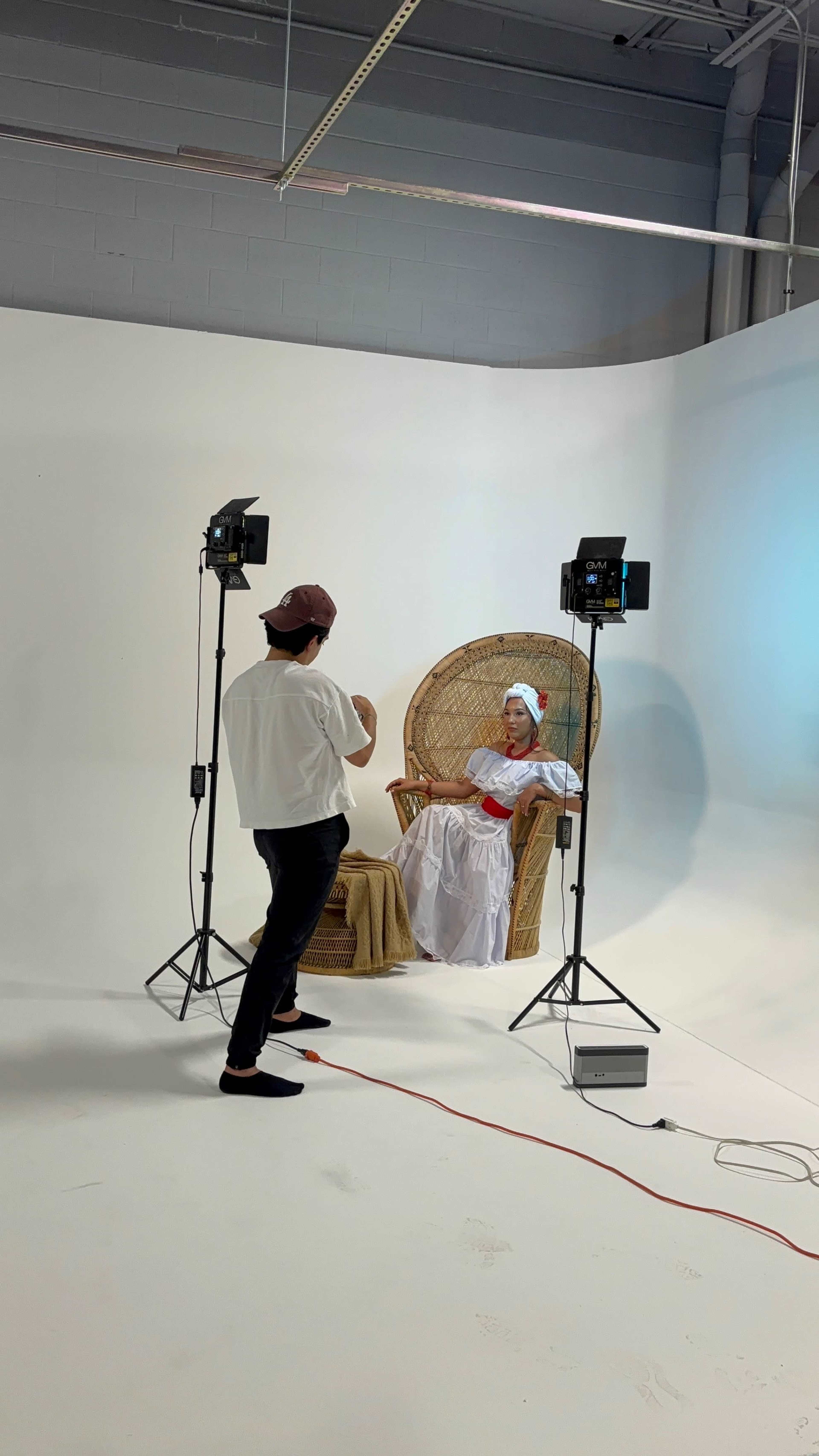 A person in a white dress with a head wrap sits in a large woven chair while another person adjusts lighting equipment in a well-lit studio.