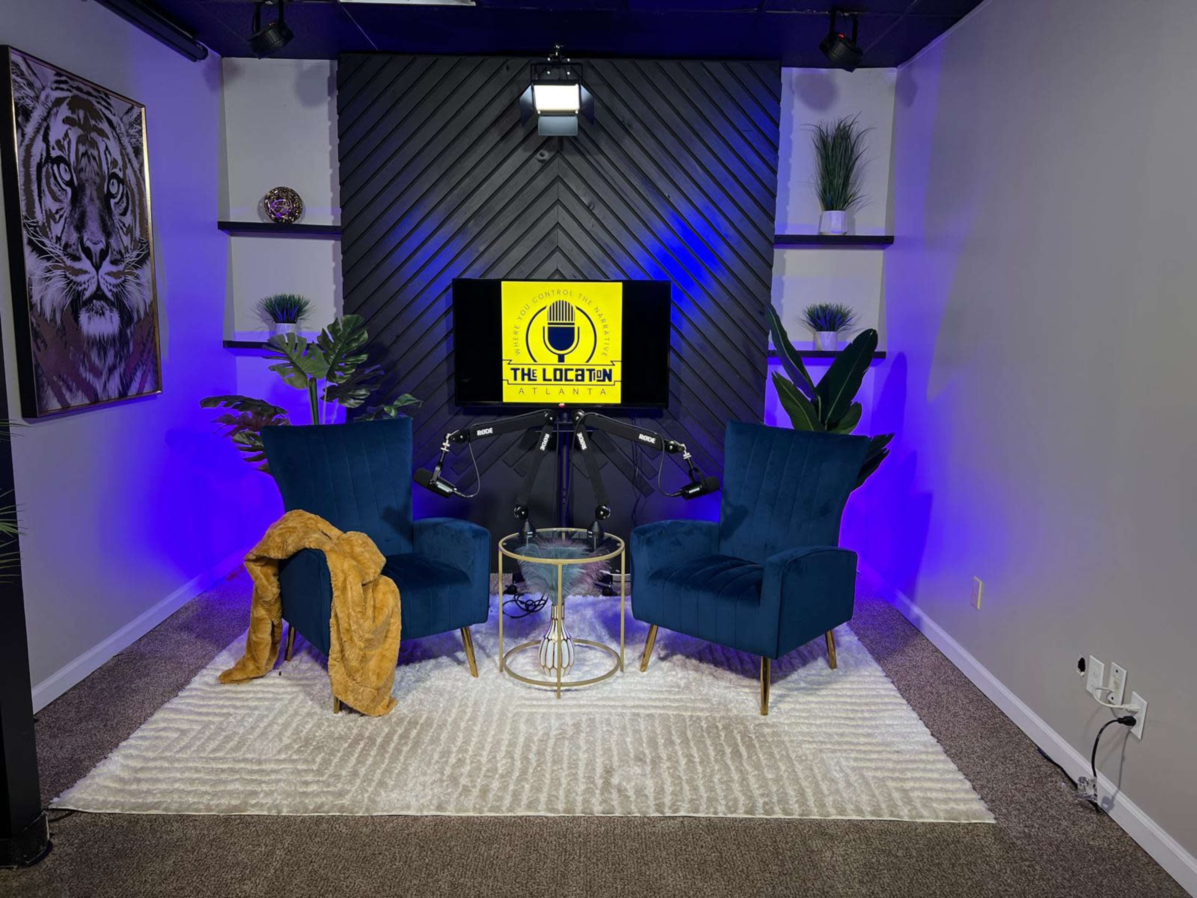 The image shows a modern studio setup with two teal chairs facing a screen displaying "THE LOCATION," surrounded by decorative shelves and a rug on the floor.