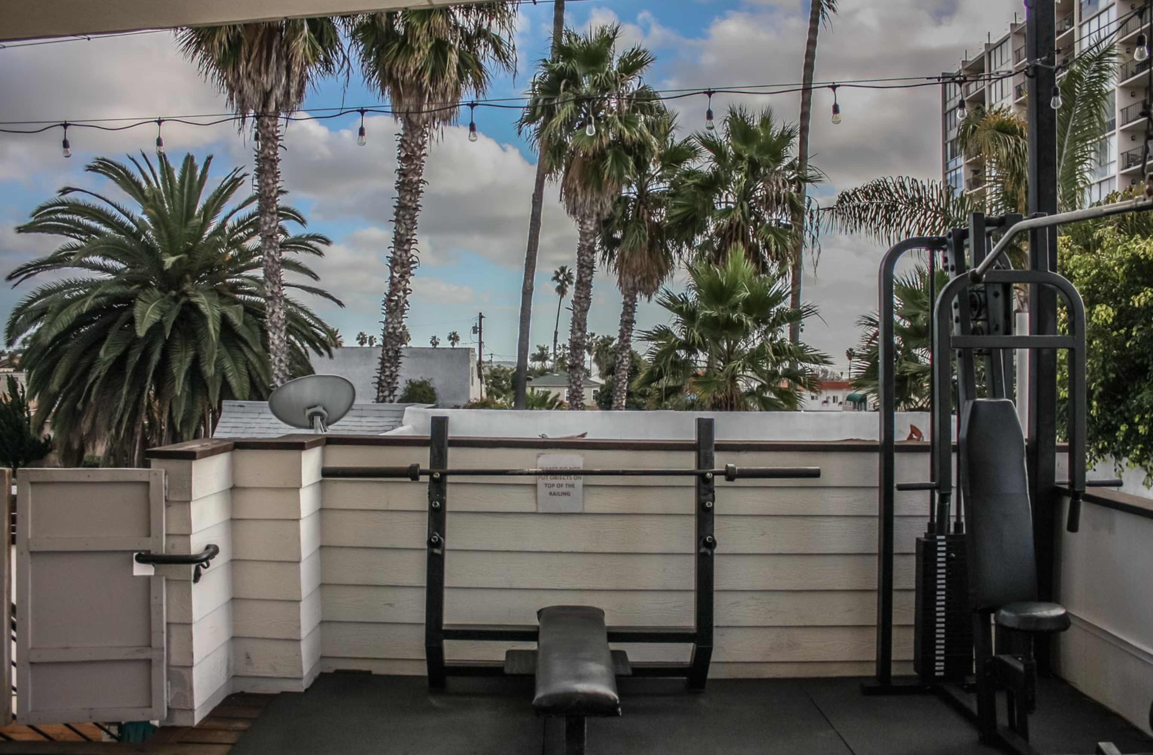 A small outdoor gym space features a weight bench and exercise equipment, surrounded by tall palm trees and a cloudy sky.