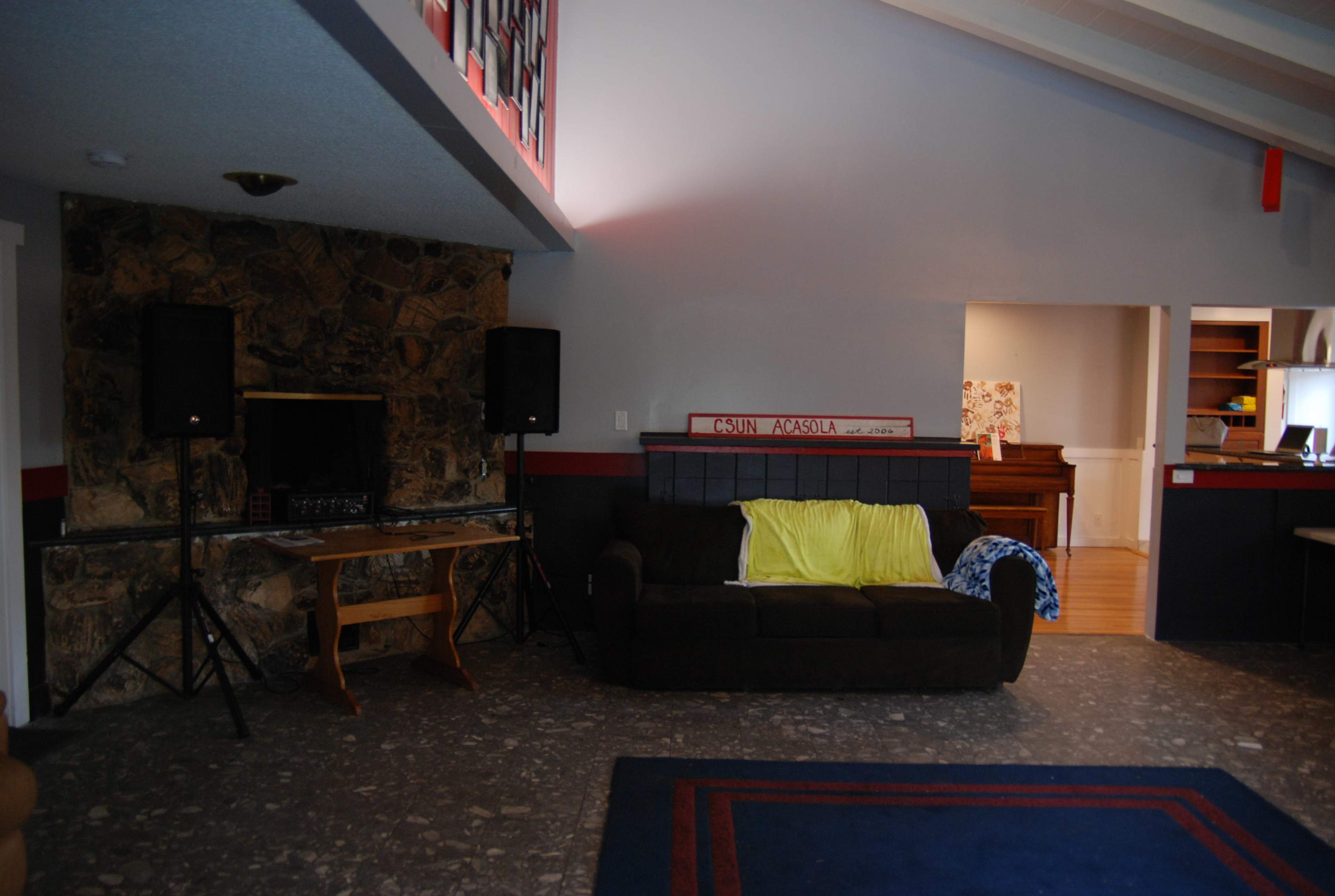 A living room featuring a green sofa, a wooden table with speakers, a stone fireplace, and a glimpse of a piano and another room in the background.