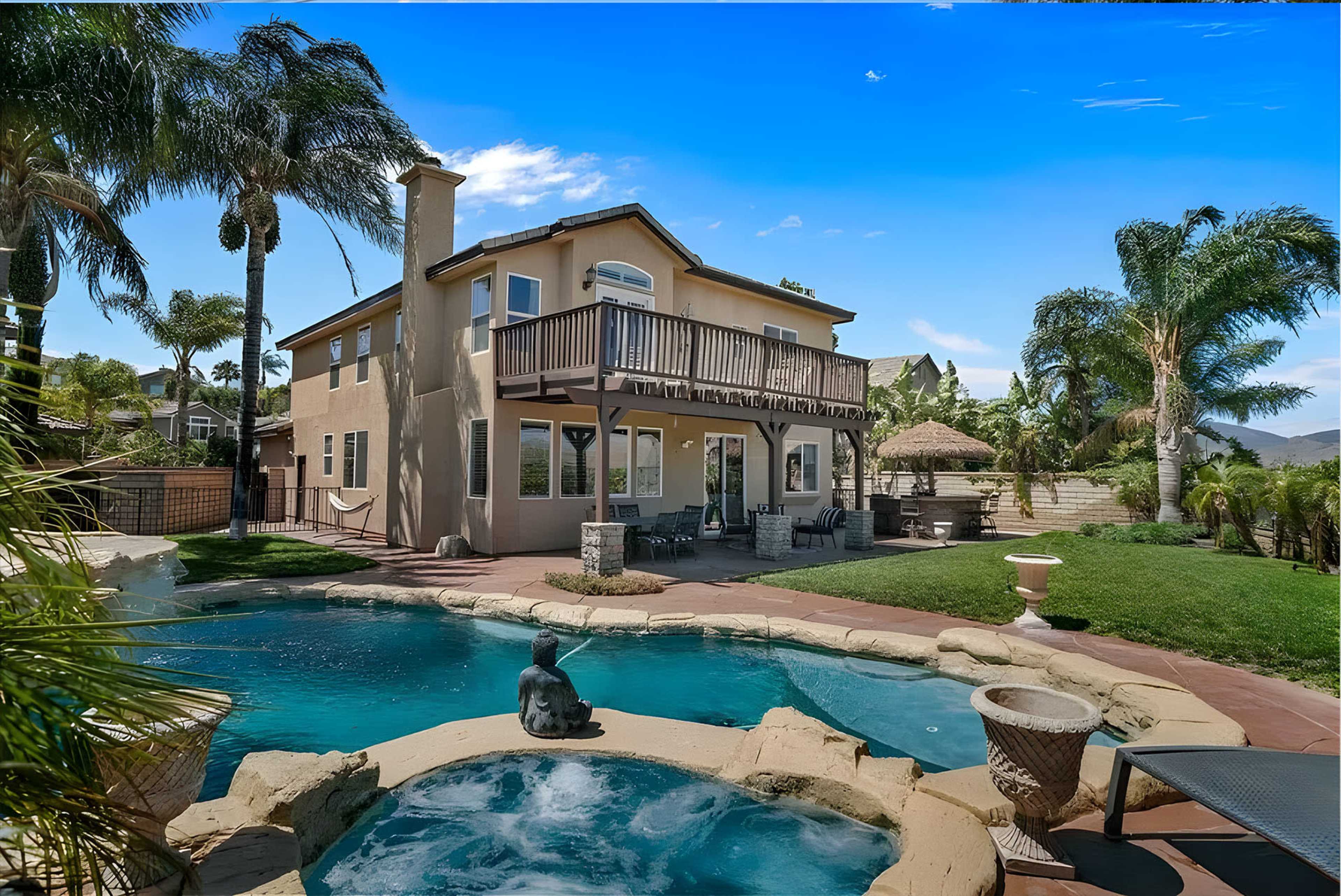 A two-story house with a balcony overlooks a pool and landscaped yard filled with palm trees and outdoor seating areas.
