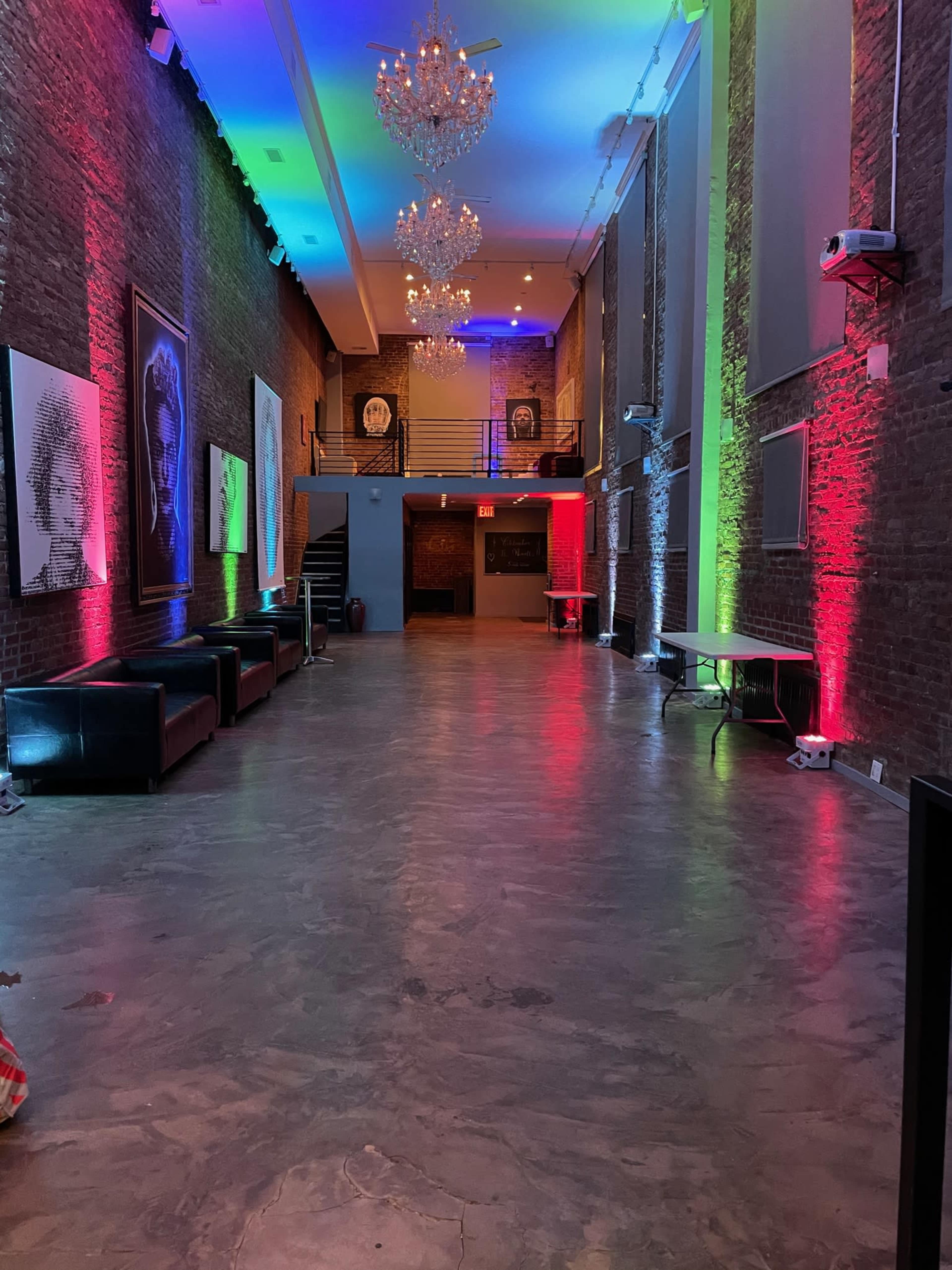A spacious event hall featuring a polished concrete floor, brick walls, and vibrant colored lighting with chandeliers hanging from the ceiling.