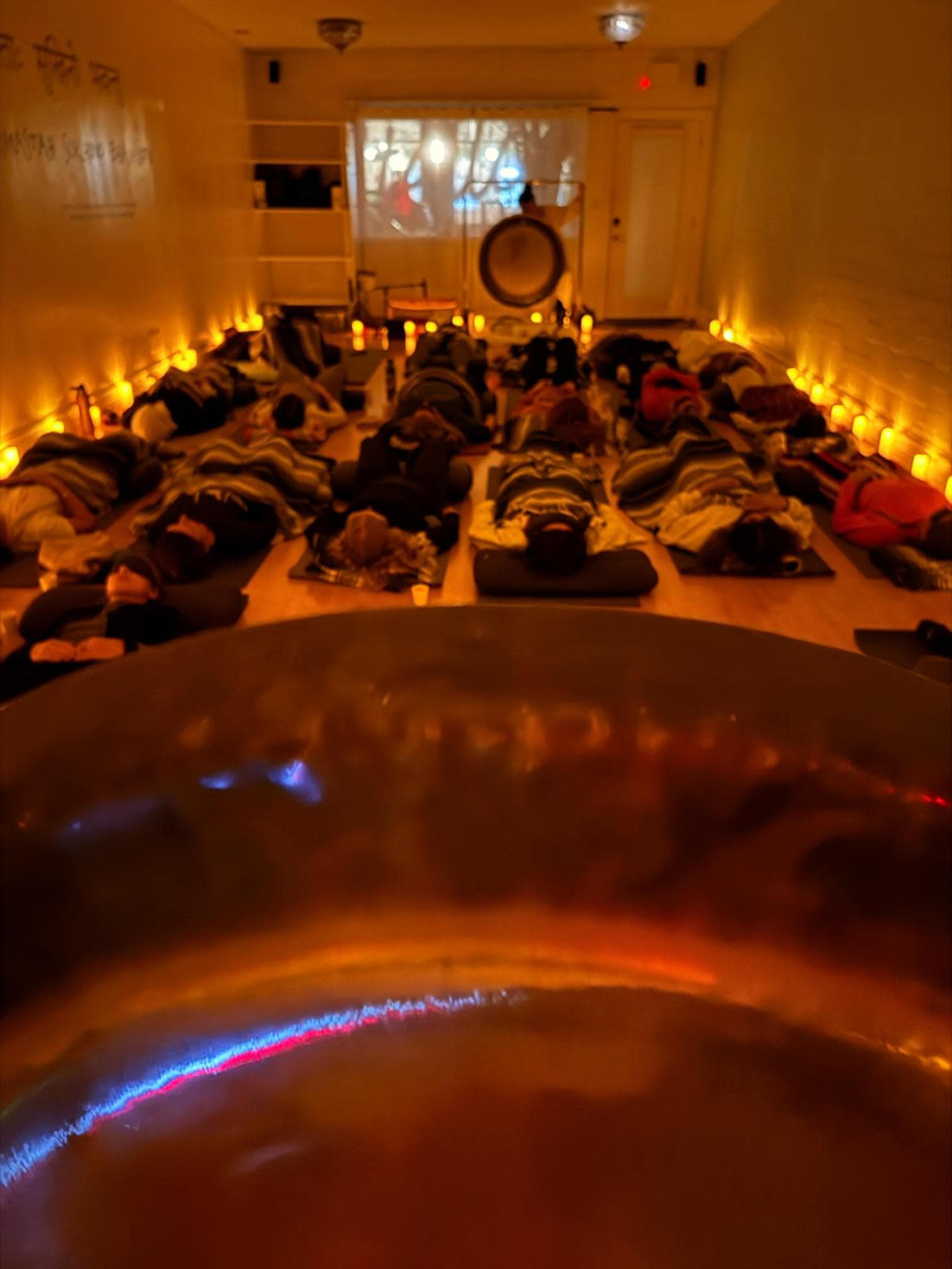 The image depicts a dimly lit room filled with people lying on mats while a large singing bowl is in the foreground, reflecting soft light.