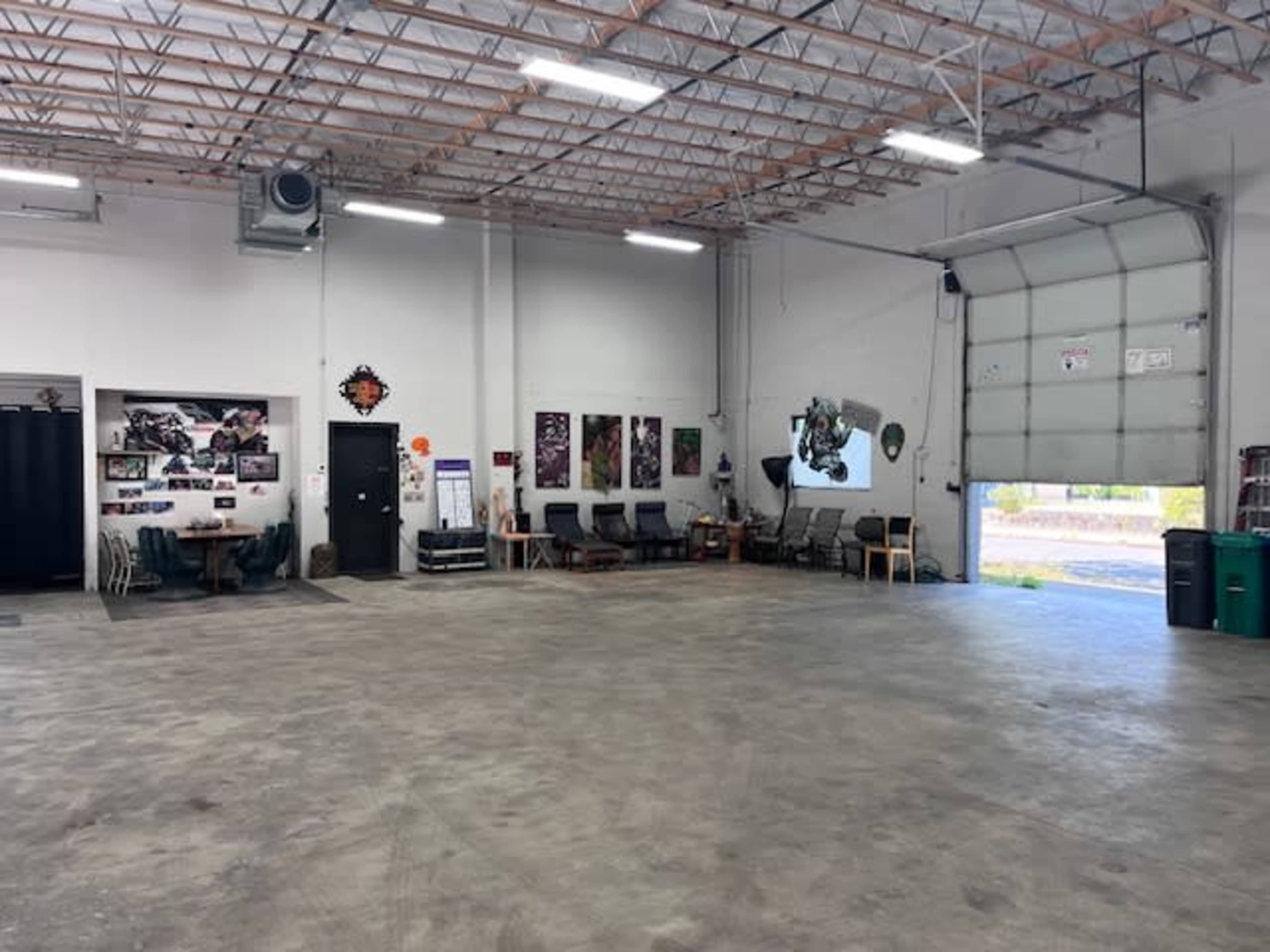 A spacious garage or workshop with unoccupied tables and chairs, wall art, and a large door that opens to an outdoor area.