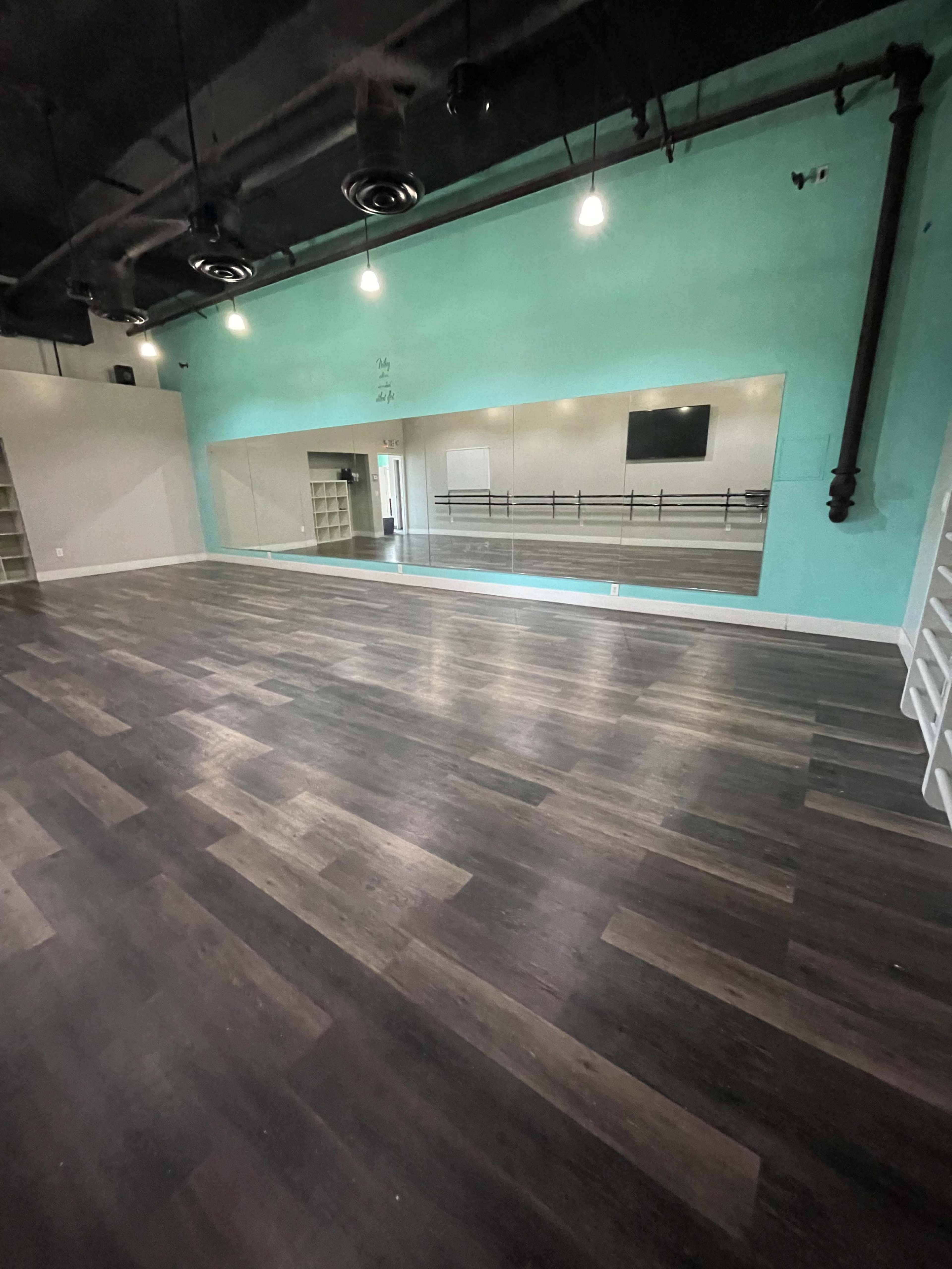 A spacious dance studio with wooden flooring, a large mirror on one wall, and teal-colored walls.