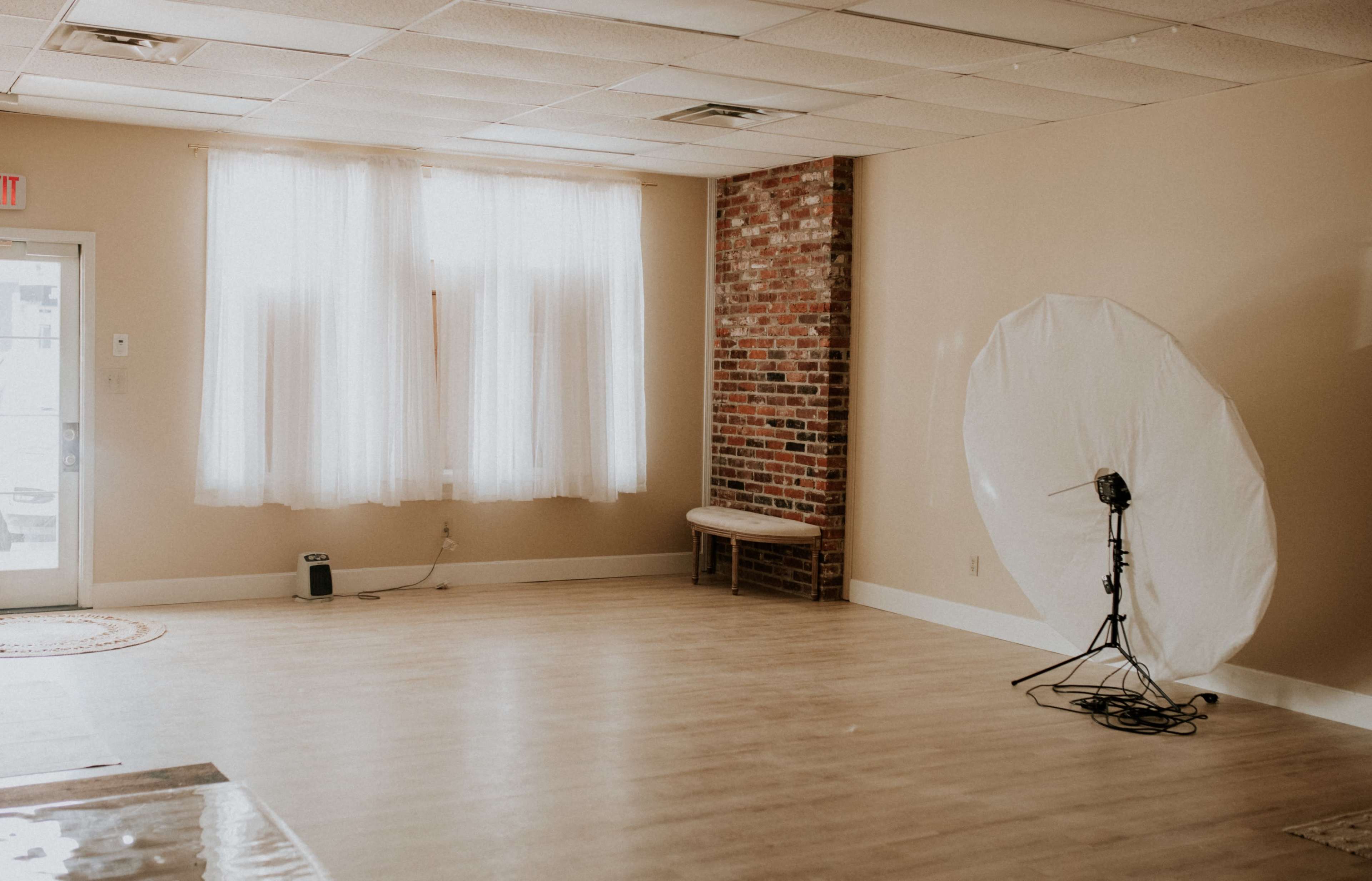 The image shows an empty room with a large white backdrop, a photography light stand, a wooden bench, and sheer curtains letting in natural light.