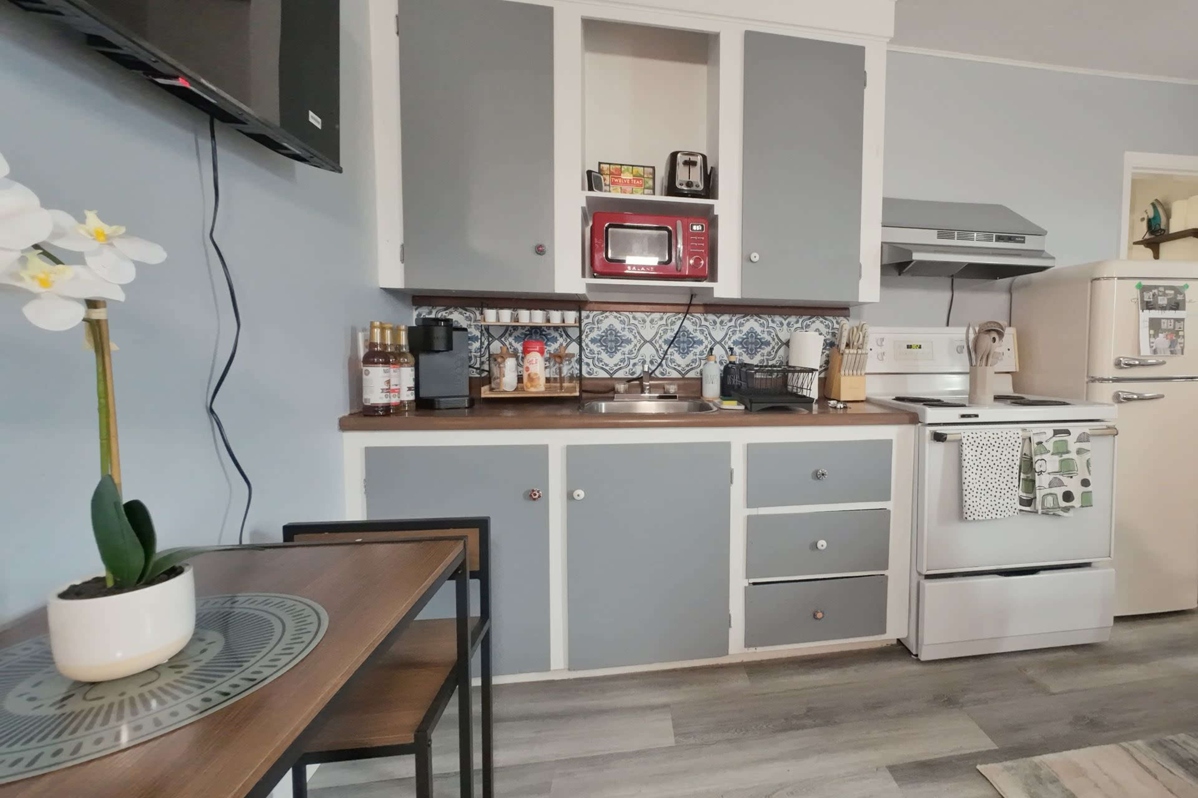 The image shows a compact kitchen with light blue walls, gray and white cabinetry, and a simple dining table.