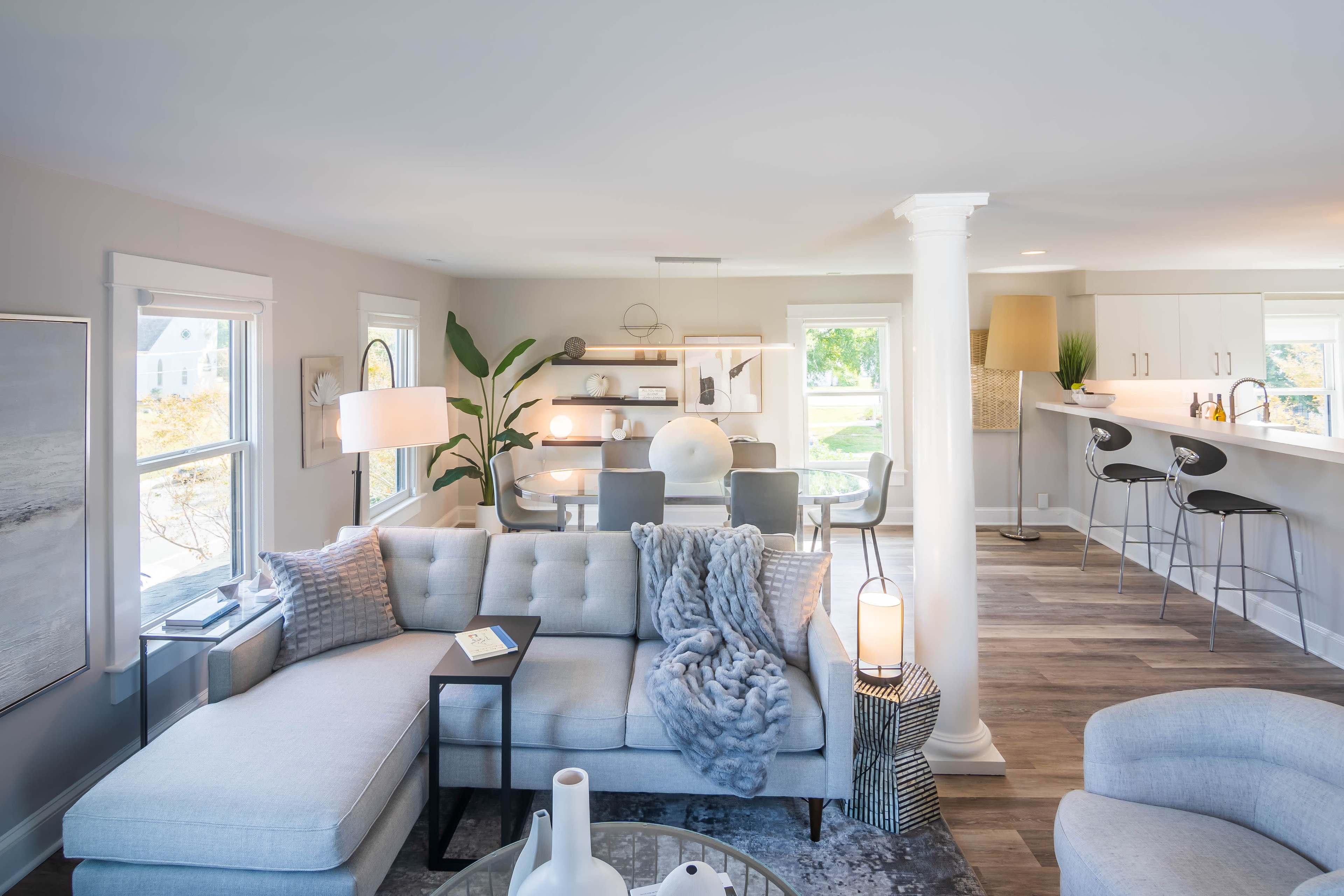 A modern living room features a light-colored sectional sofa, a coffee table, and a dining area with bar stools in the background.