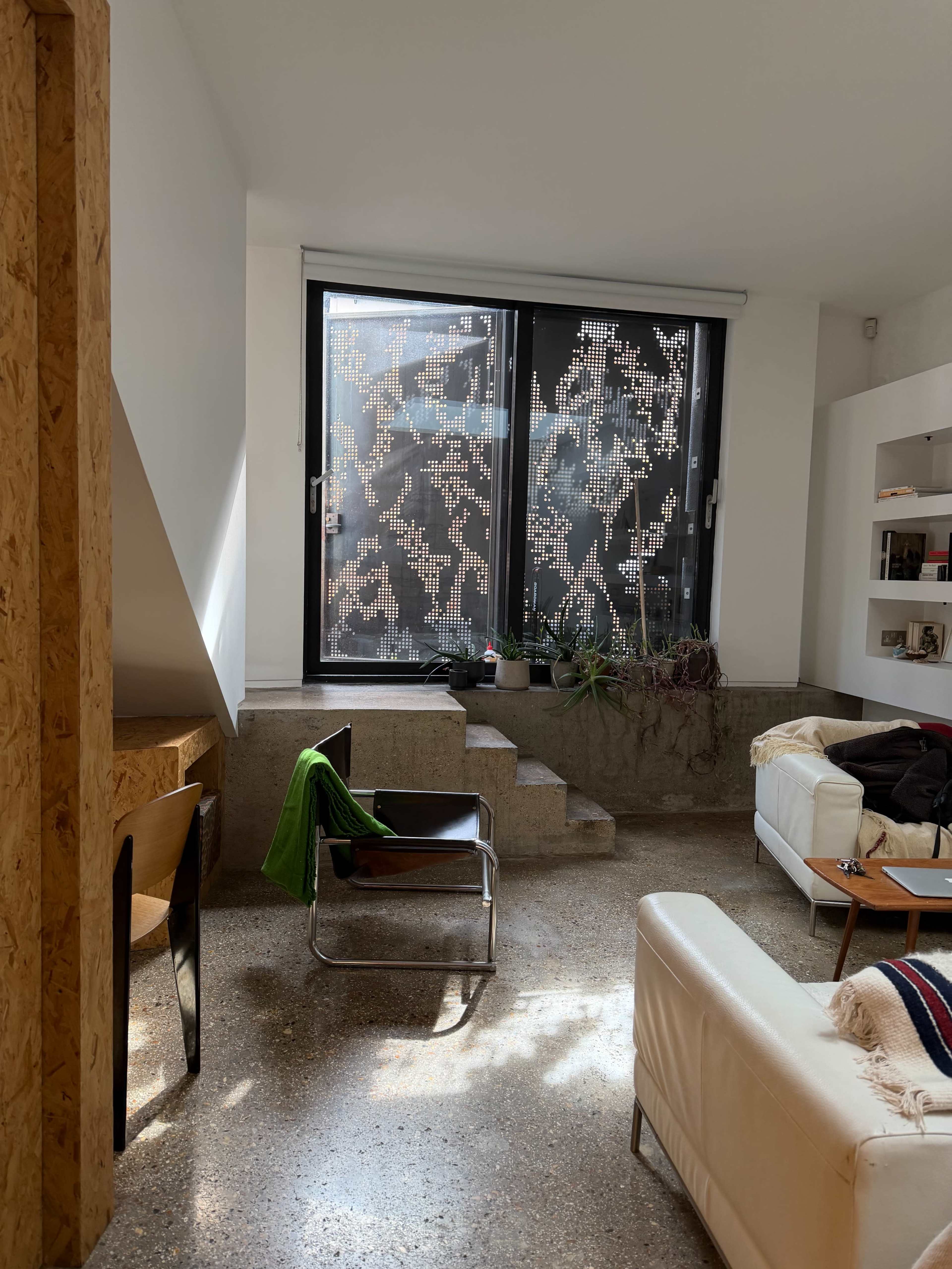 A modern living space features a white couch, a simple chair, and a large window with patterned glass, allowing light to illuminate the room.