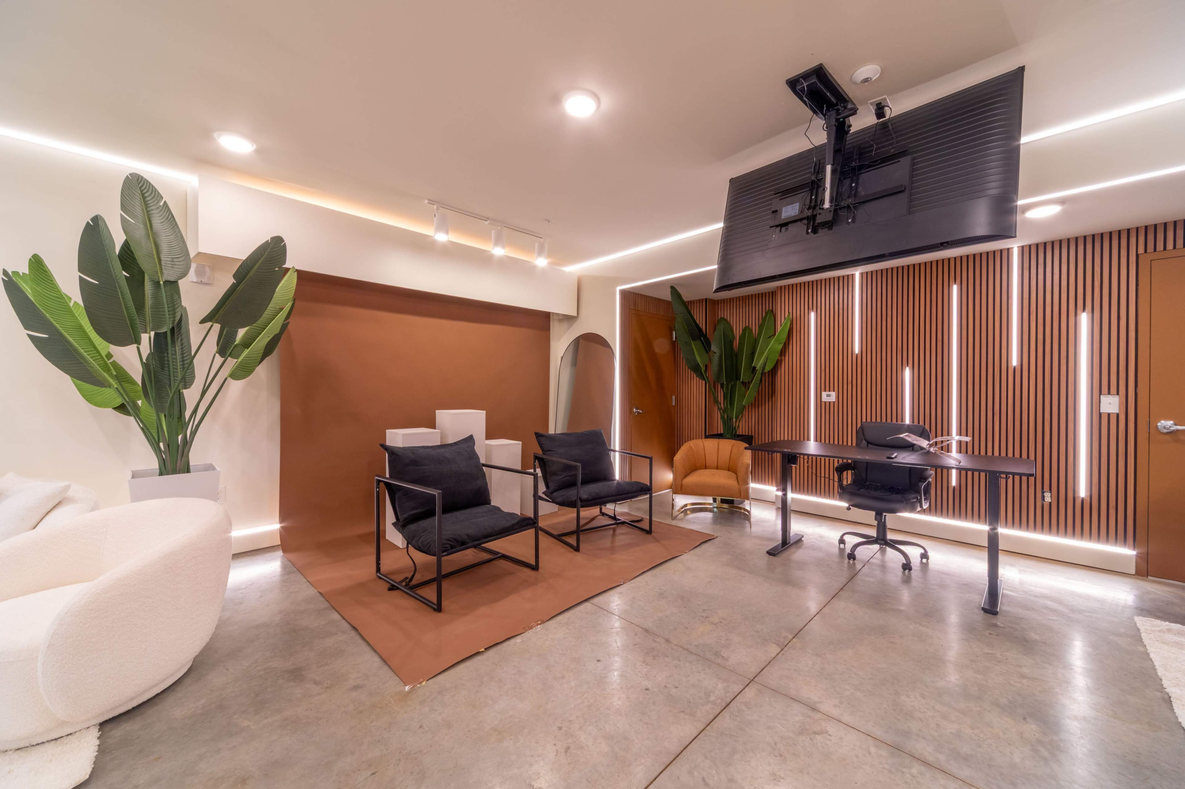 The image shows a modern interior space with two black chairs and a desk against a brown wall backdrop, flanked by plants and illuminated by recessed lighting.