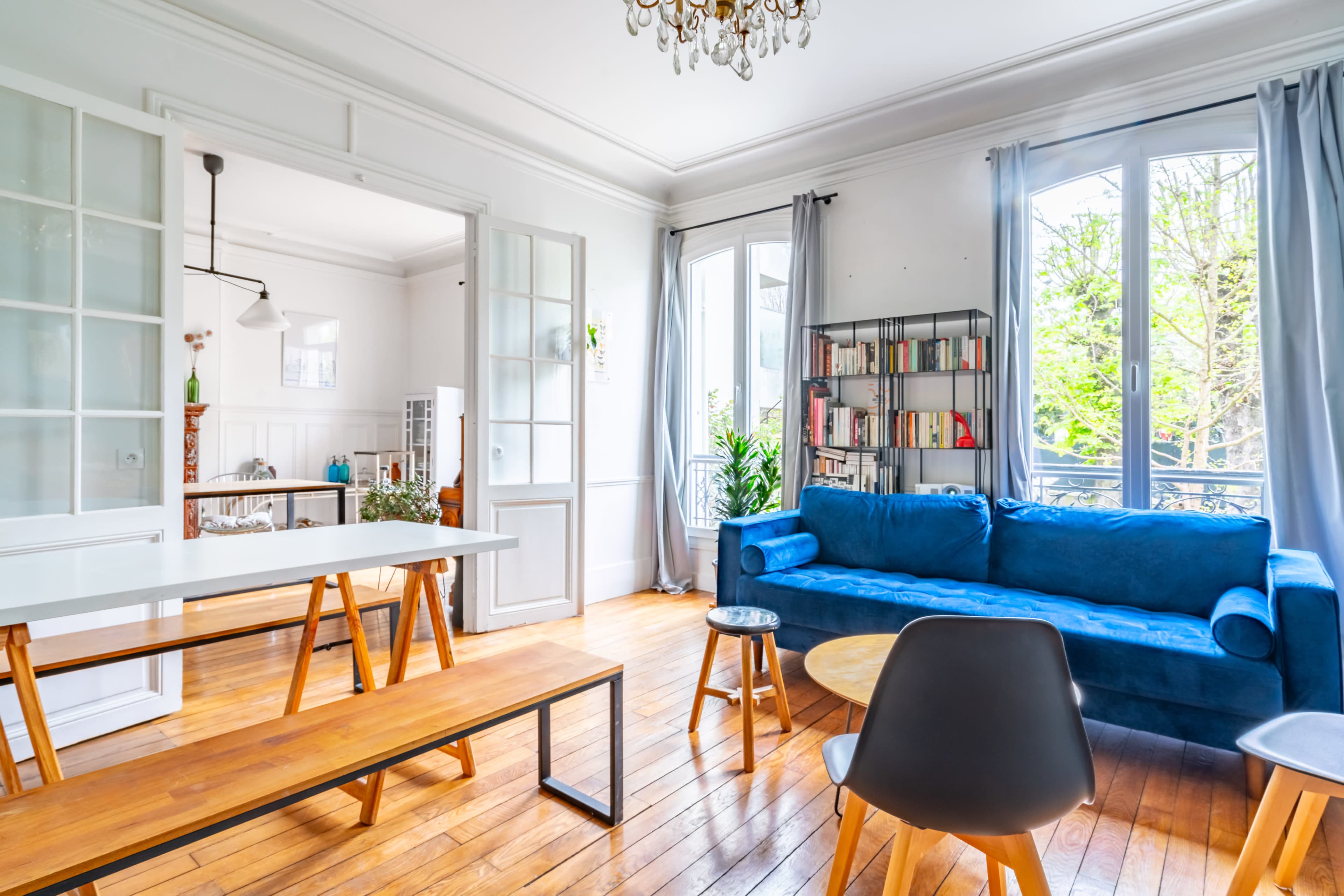 A bright living room features a blue sofa, wooden furniture, and large windows that let in natural light.
