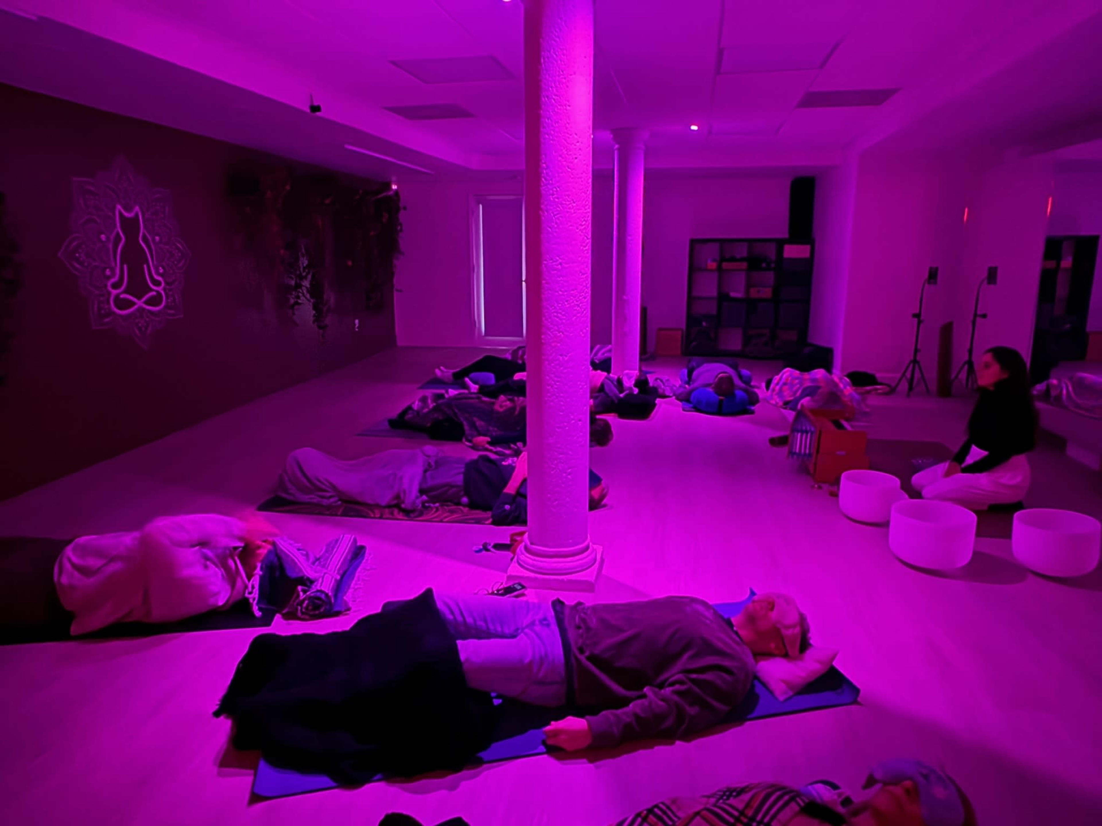 A group of people lie on mats in a dimly lit, pink-hued room designed for relaxation or meditation.