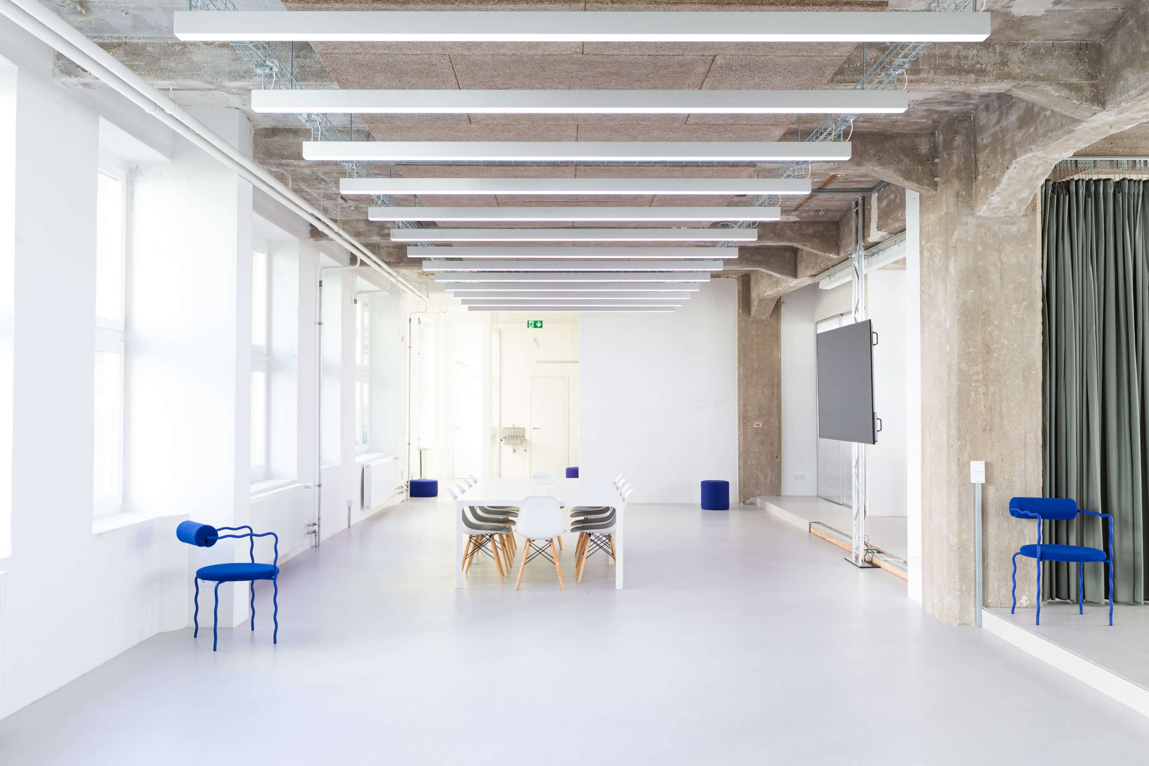 The image shows a spacious, modern room with a long table and chairs, large windows, concrete walls, and overhead lighting.