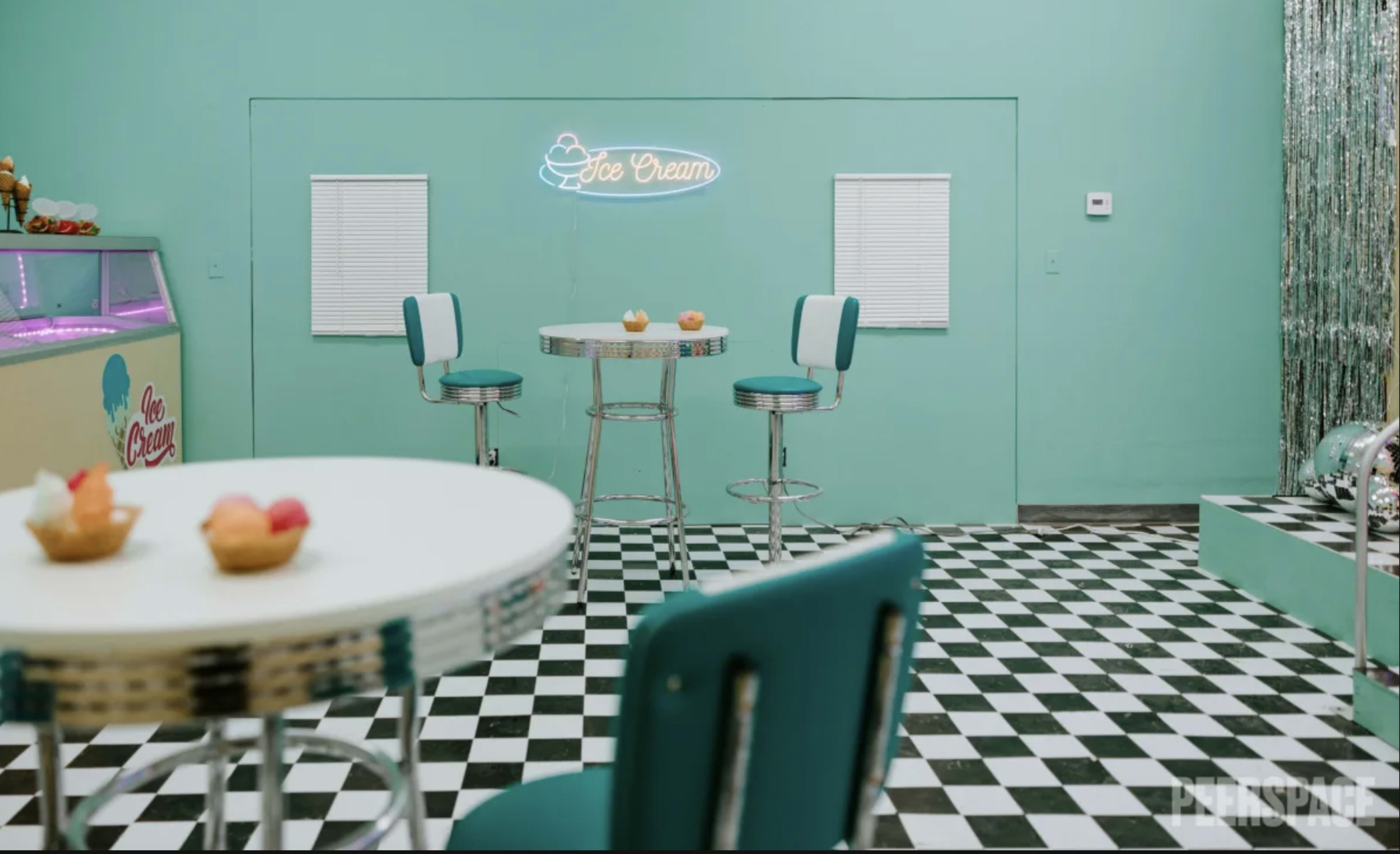A pastel-colored ice cream parlor with two round tables, four stools, and a neon "Ice Cream" sign on the wall.