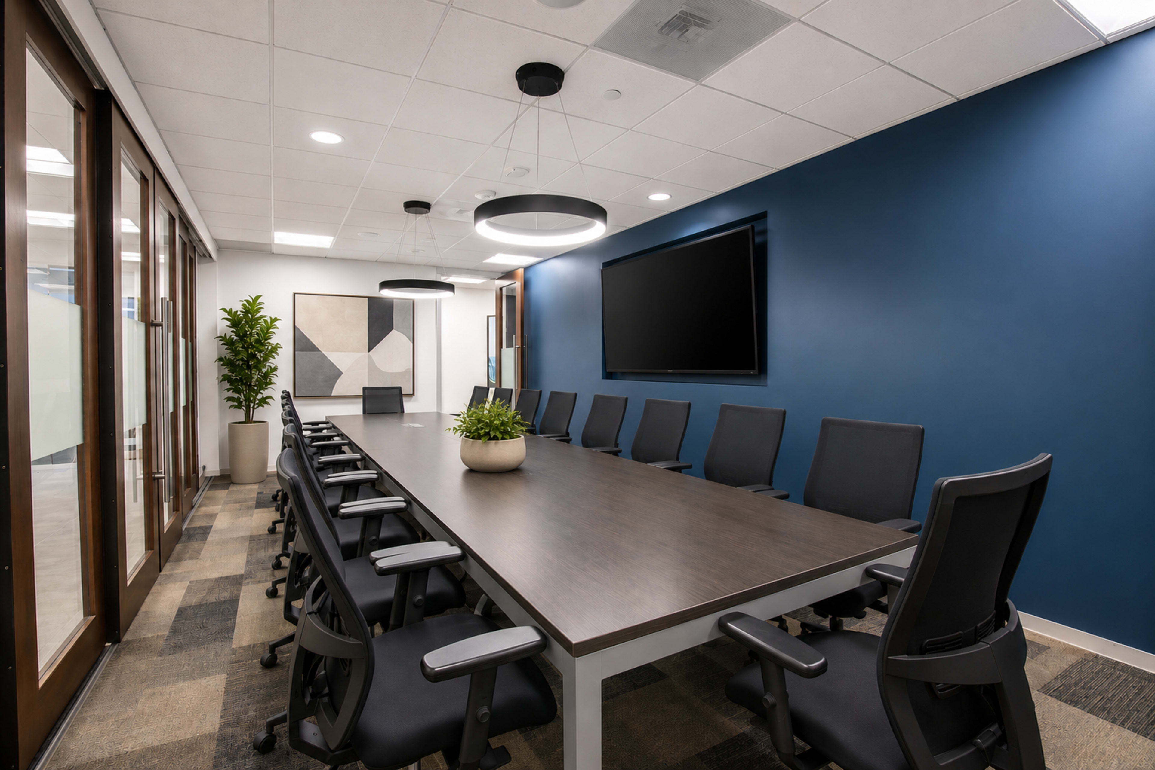 A modern conference room features a large wooden table surrounded by black swivel chairs, with a TV mounted on a blue wall and a potted plant in the center.