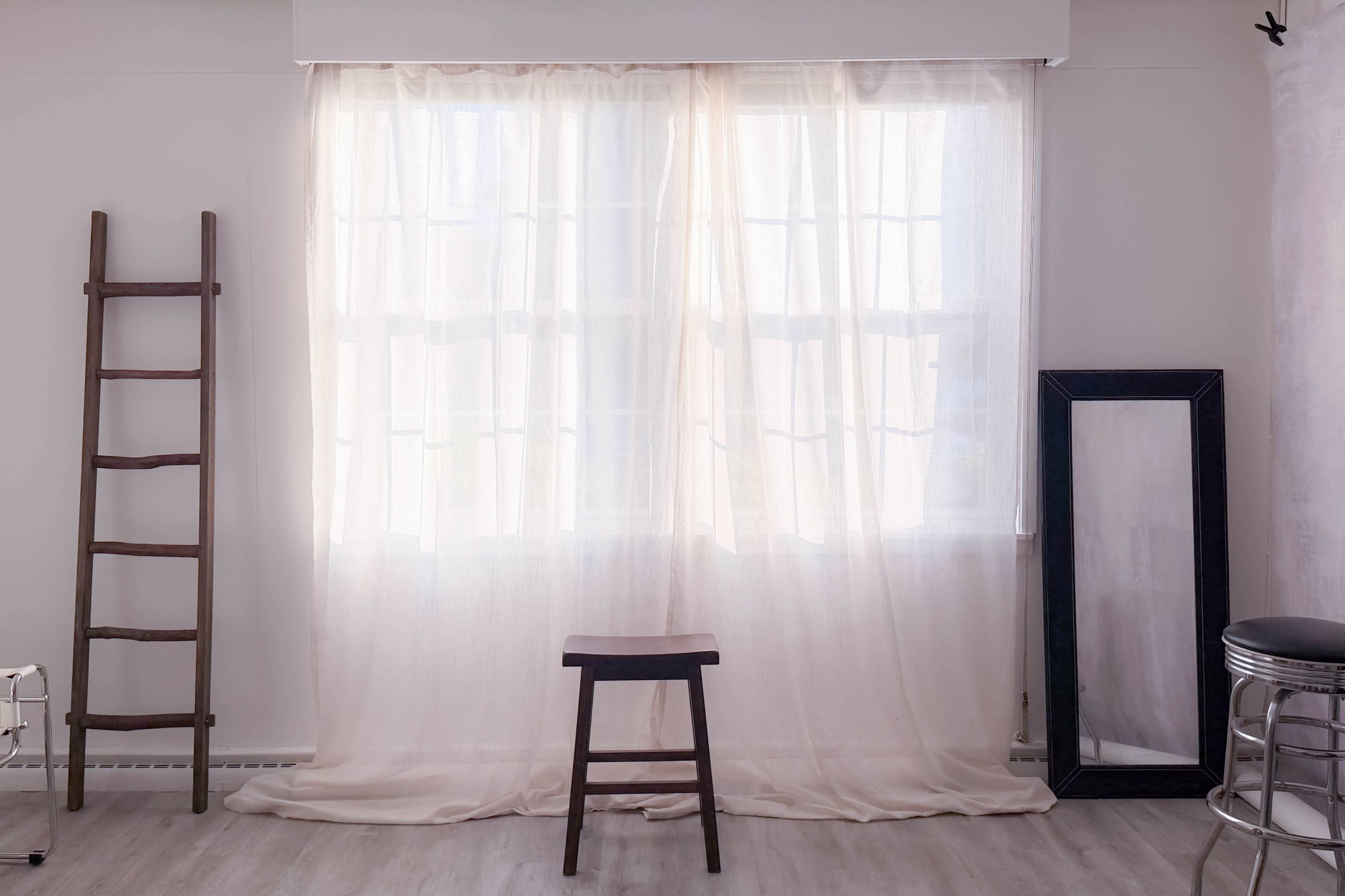 A wooden ladder stands against a white wall beside a mirror, while a stool is positioned in front of sheer curtains covering a window.