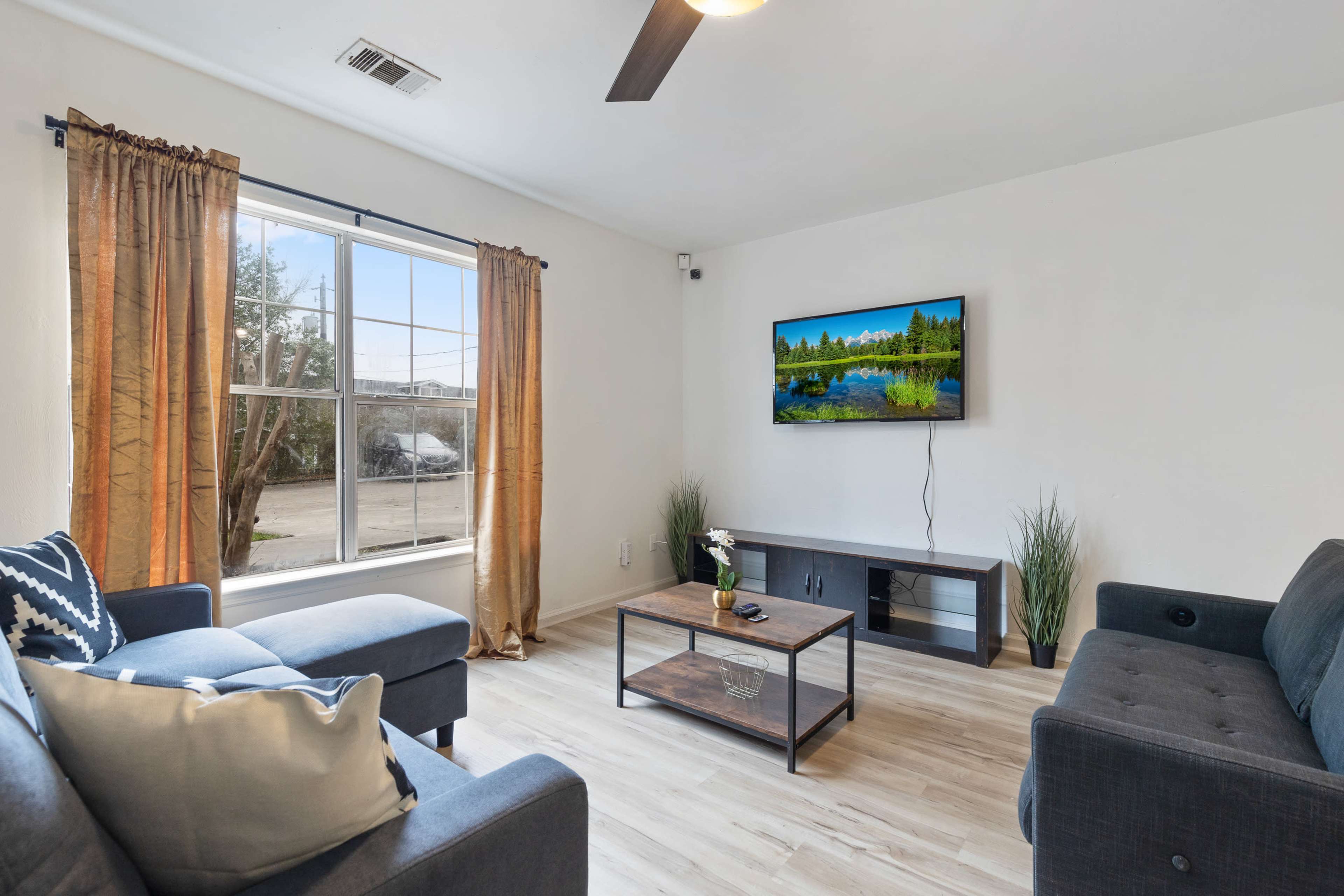 The living room features two gray sofas, a coffee table, a television mounted on the wall, and large windows with light curtains.
