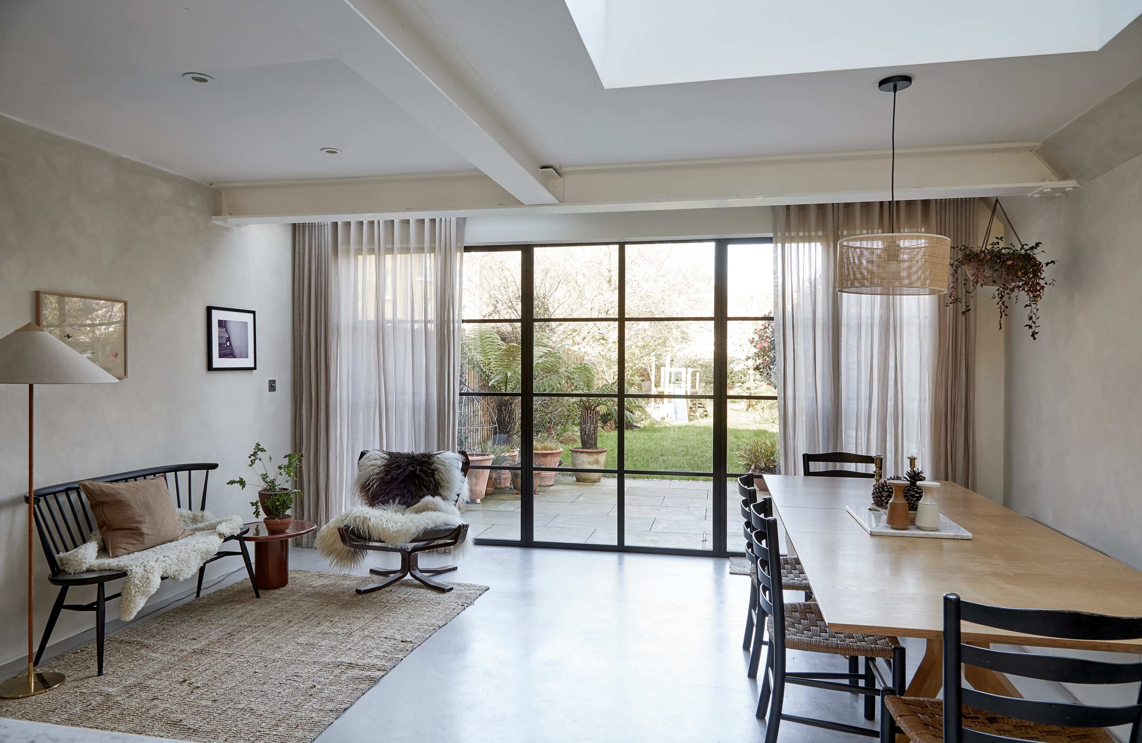 The image shows a modern dining area with a large table, black chairs, a minimalist lamp, and a view of a garden through sliding glass doors.